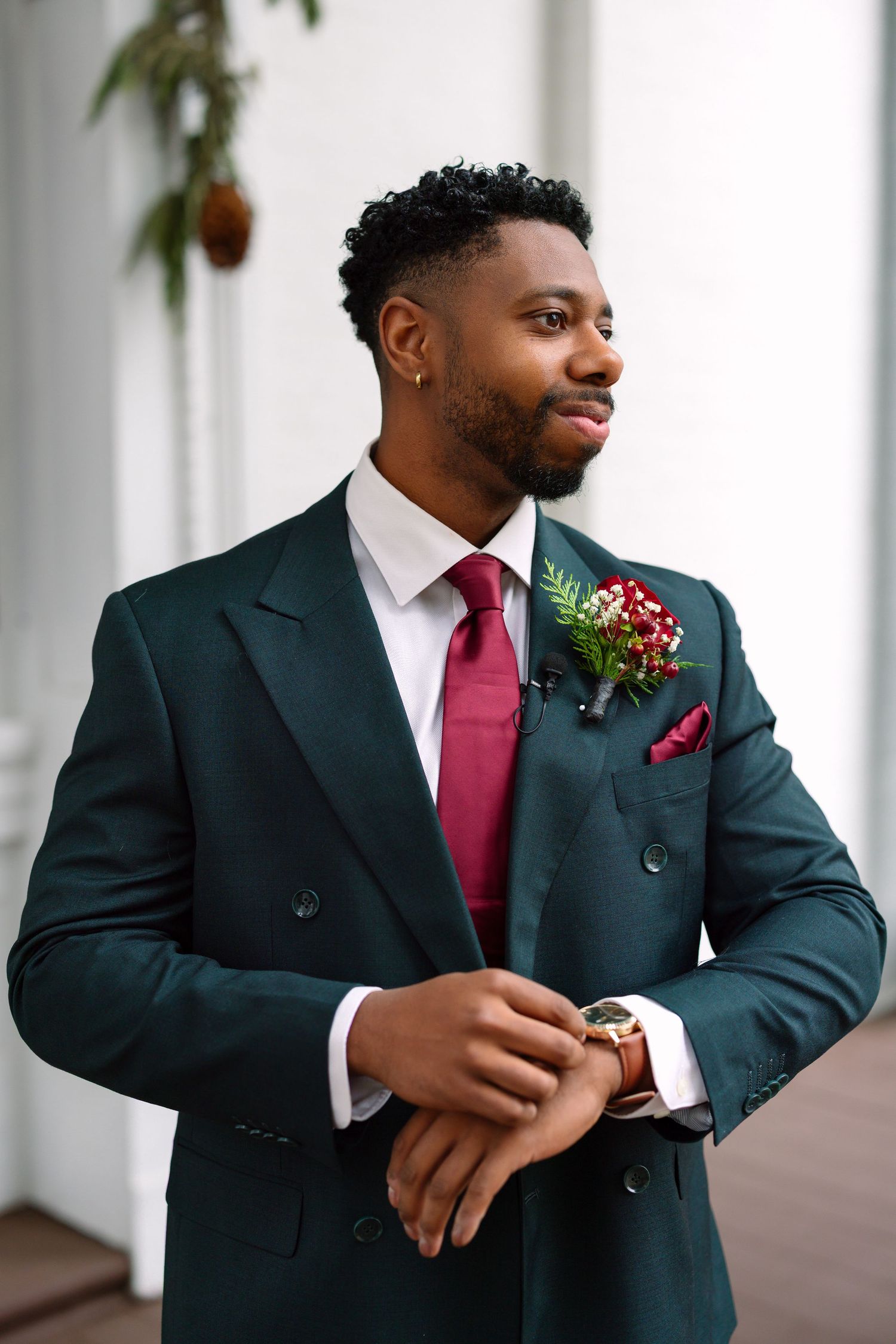 Dapper groom in dark green double-breasted suit with burgundy tie and floral boutonniere adjusts cufflinks.