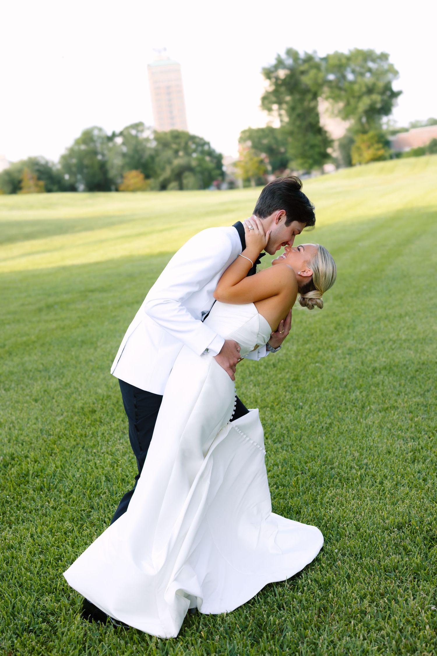 Groom in white jacket dips and kisses bride in white gown on lush green golf course lawn.