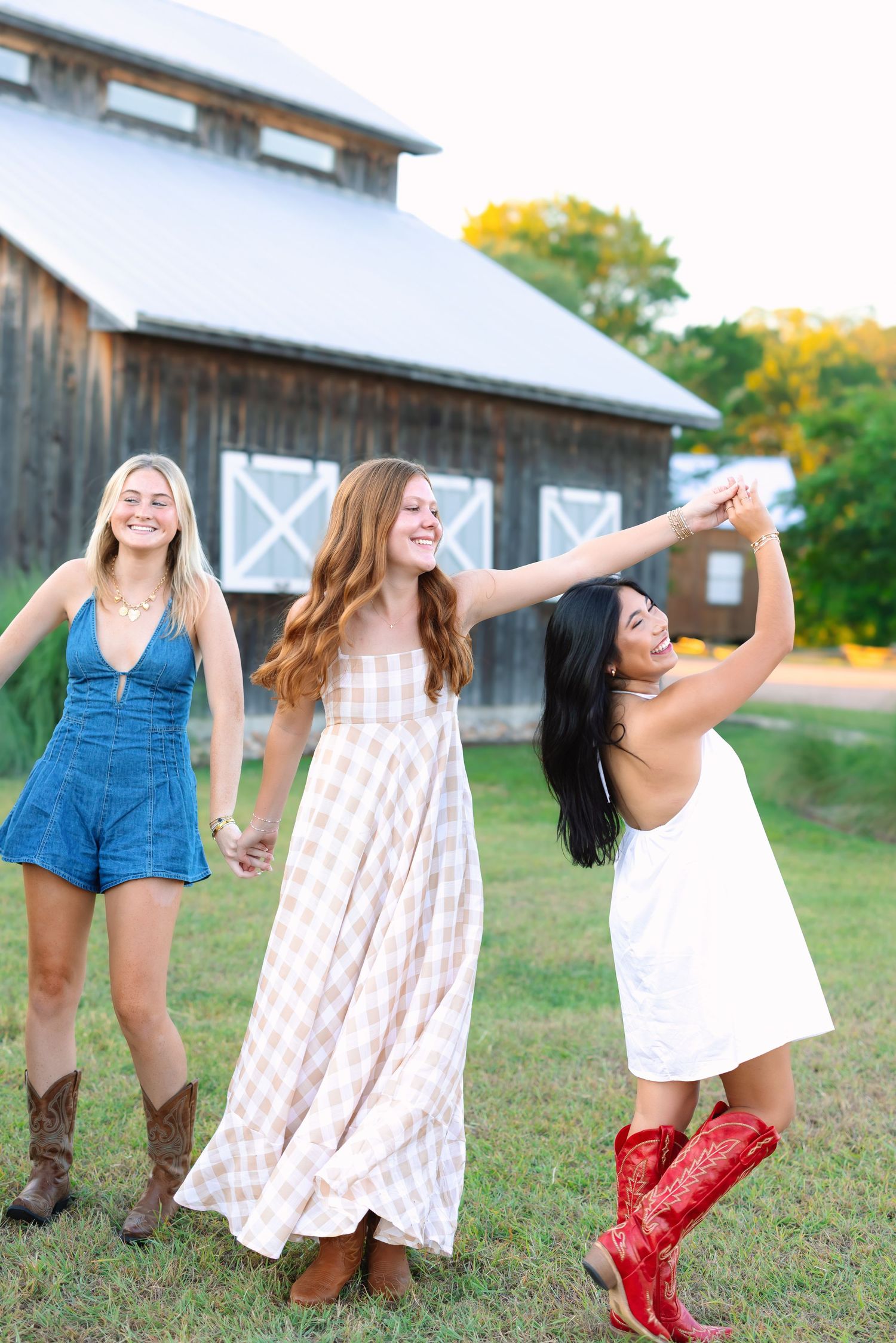 Three friends in summer outfits and boots dancing playfully by a barn.