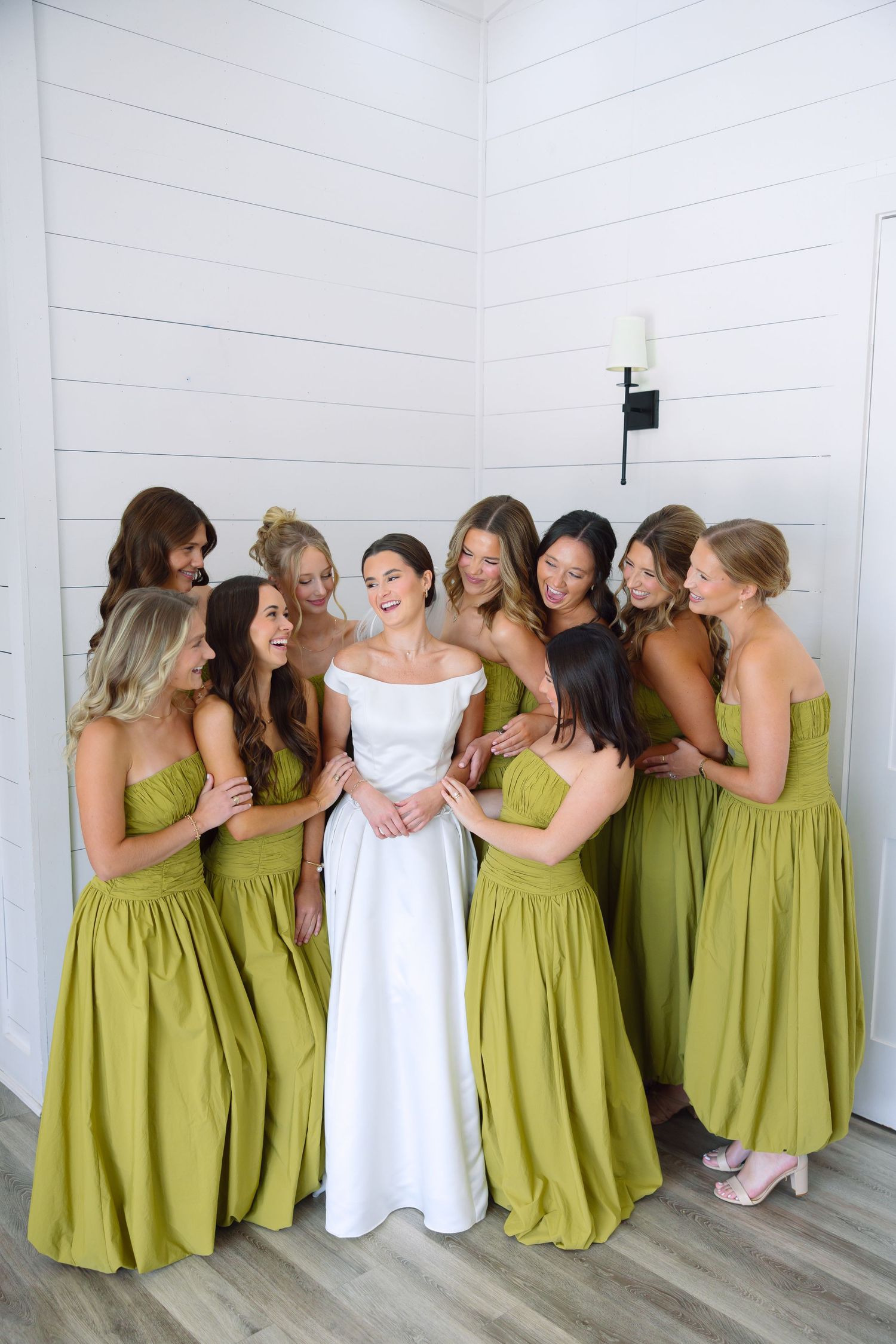 Bride in white off-shoulder gown surrounded by eight bridesmaids in olive green dresses laughing together indoors.