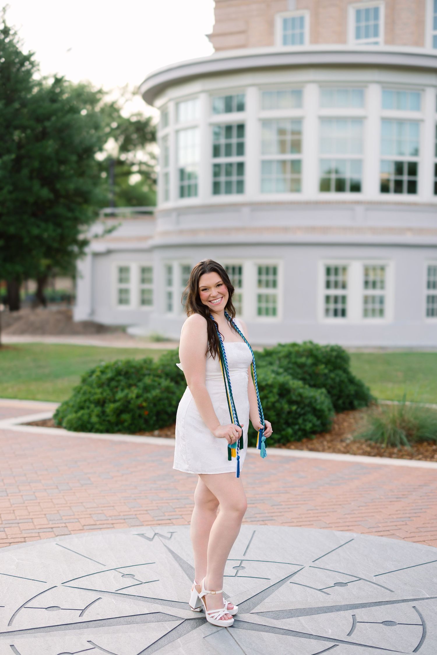 Smiling graduate in white dress holding colorful cords stands on compass rose outside university building.