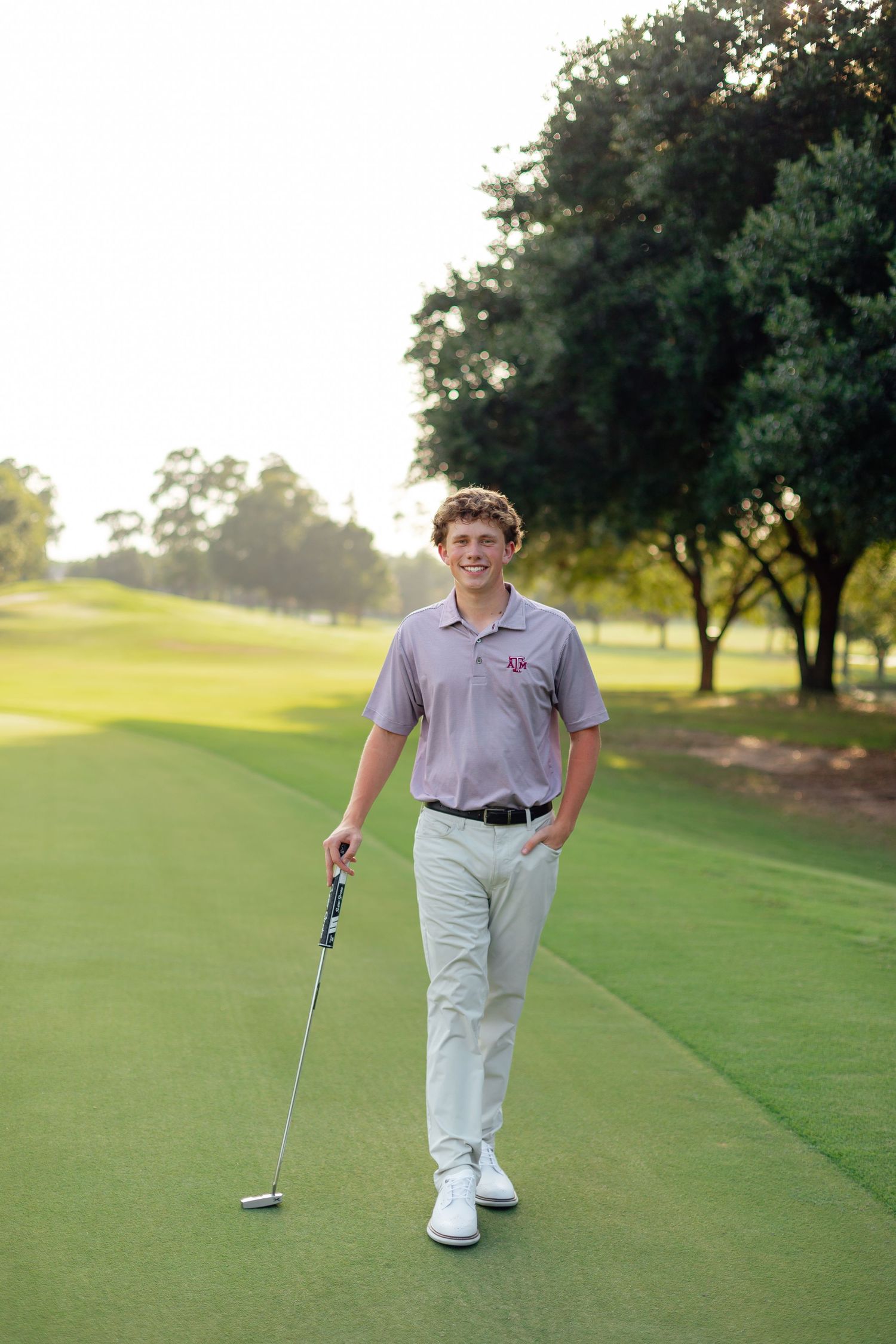Golfer practicing putting stance on manicured green with trees in background.