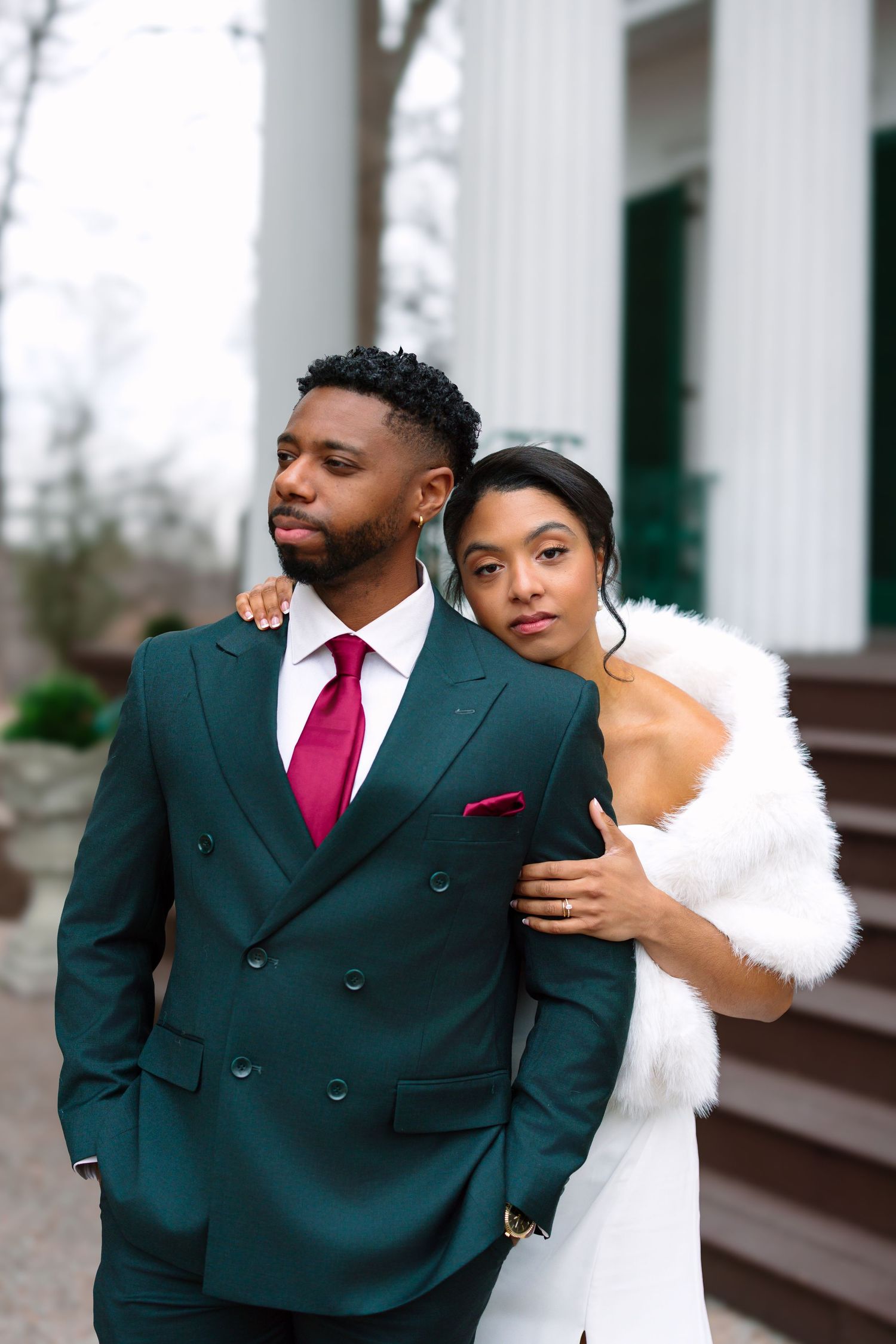 Elegant couple in green suit with red tie and white fur stole posing outside a white building.
