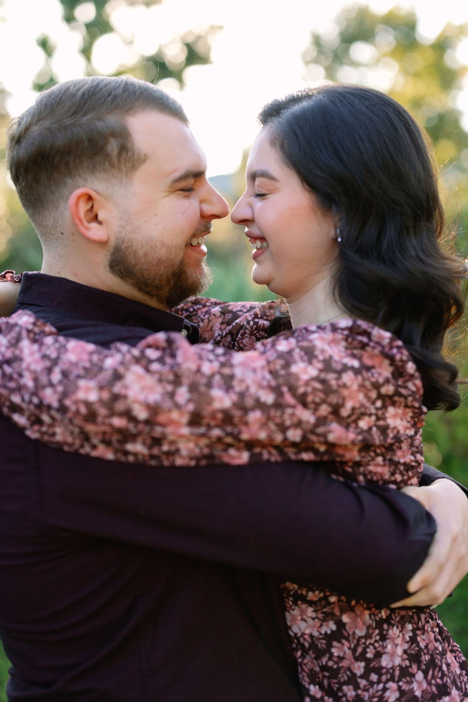 A happy couple embraces and laughs together outdoors, the woman in a floral dress and the man in a dark shirt.