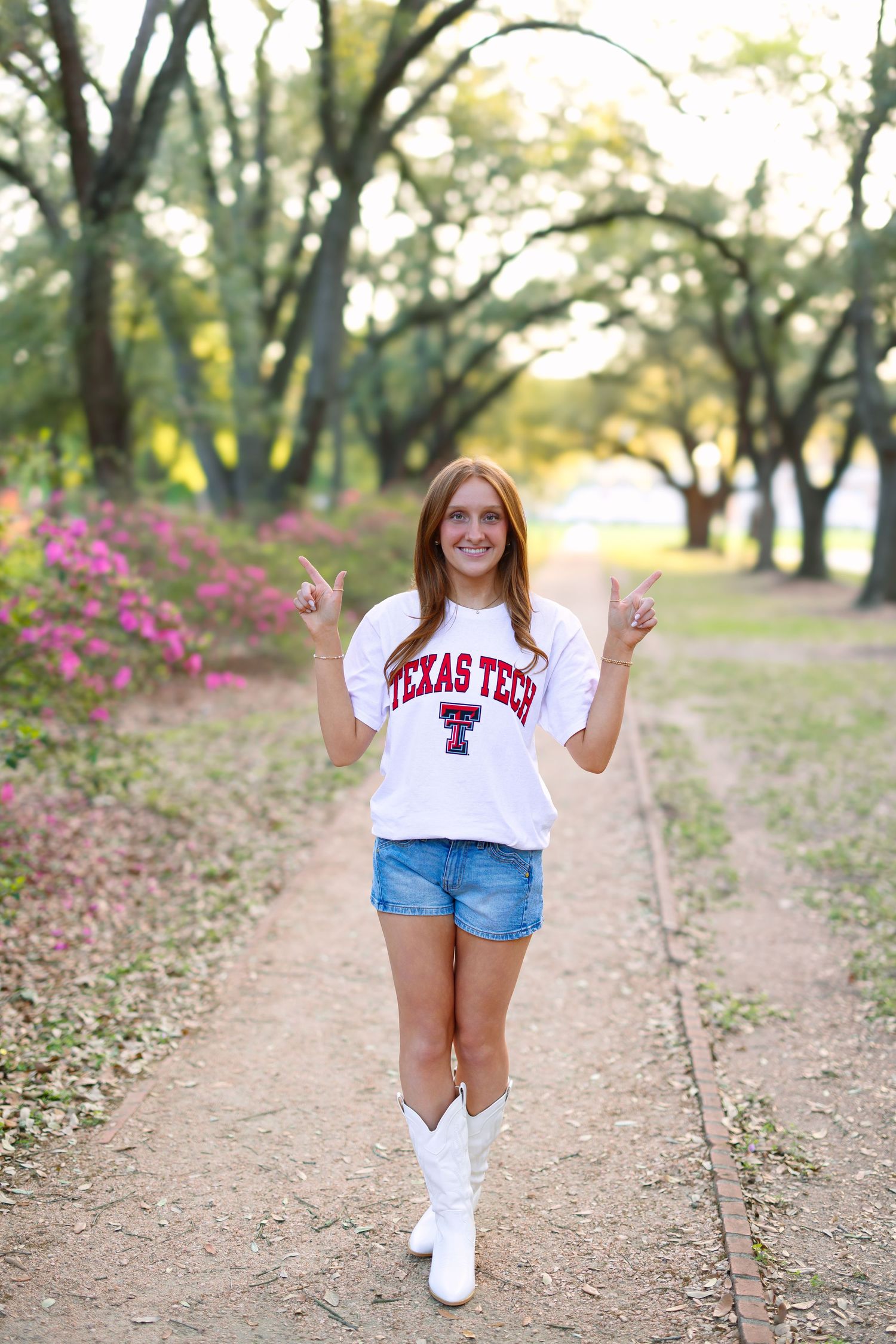 Young woman in Texas Tech shirt and white boots giving gun hand signs on a tree-lined path.