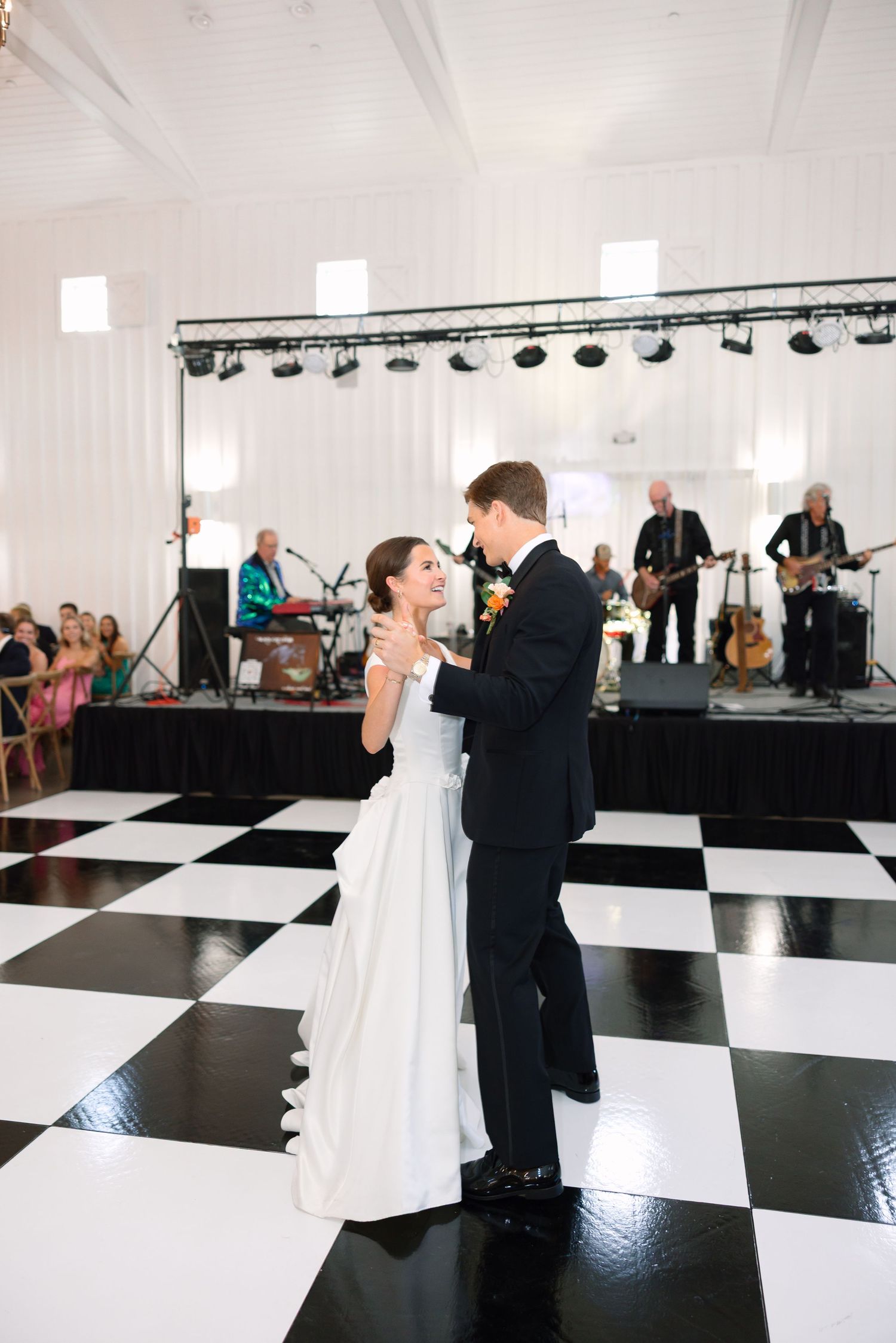 Bride and groom share first dance on checkered floor with live band performing at elegant wedding reception.