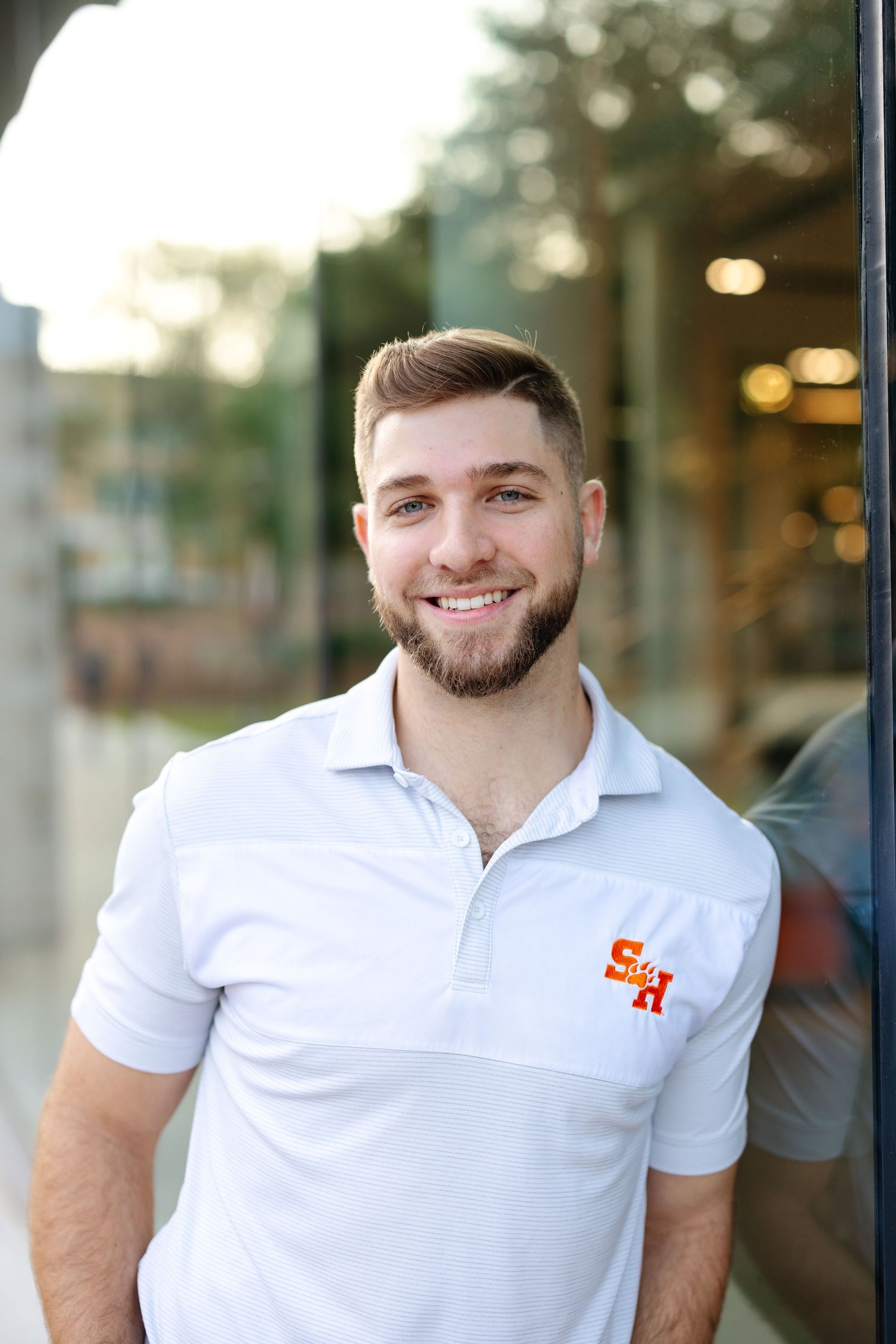 Outdoor portrait in white polo shirt with orange logo against blurred urban background.