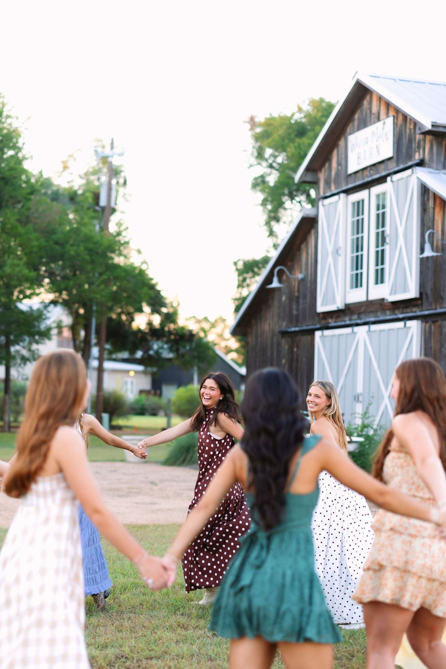 A group of friends dance in a line while holding hands in front of a rustic barn.