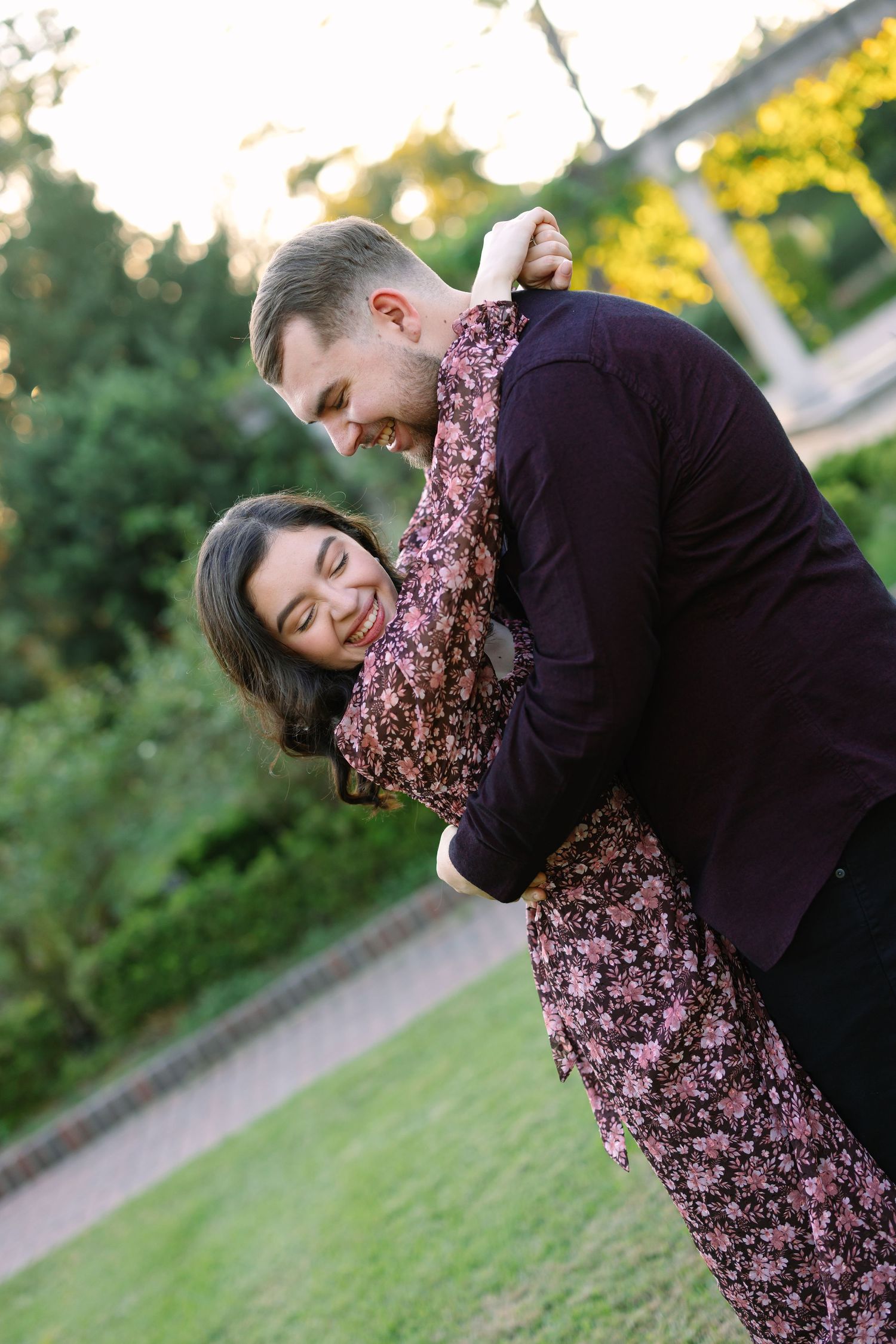 Happy couple embracing and laughing together in a sunlit park, woman in floral dress hugging man in dark blazer.