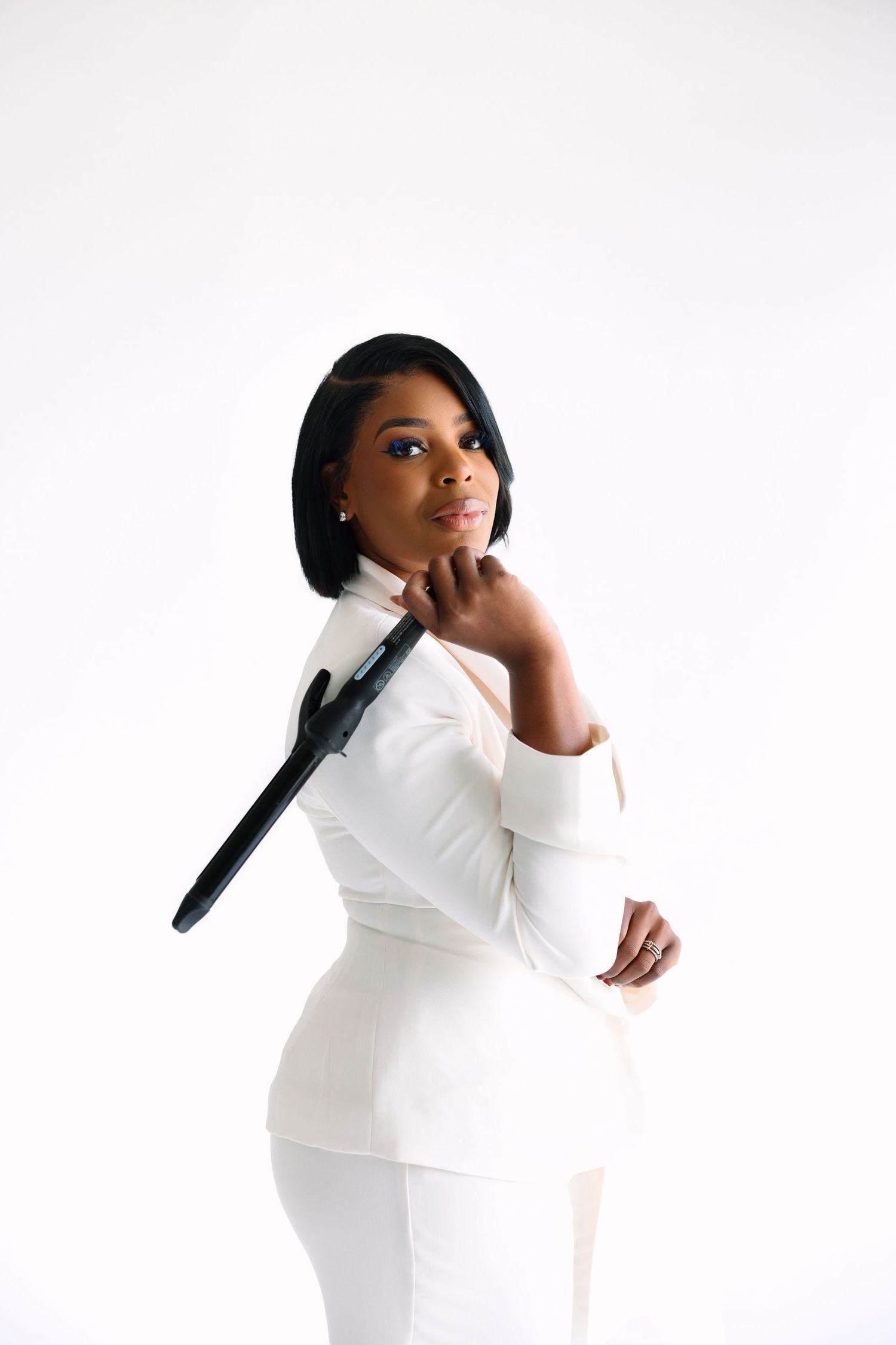 A woman in a white fitted outfit holds a curling iron over her shoulder against a white background.