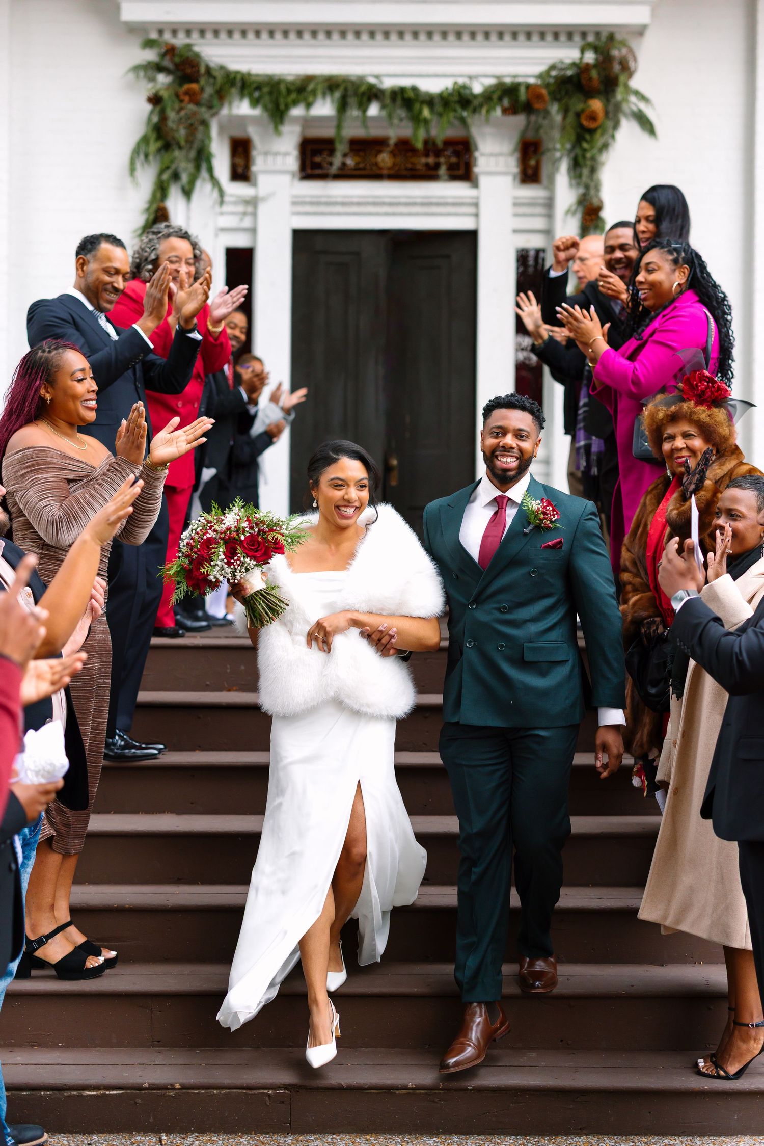 Joyful newlyweds exit church steps as guests applaud, bride in white gown with fur wrap holding red bouquet, groom in green suit.