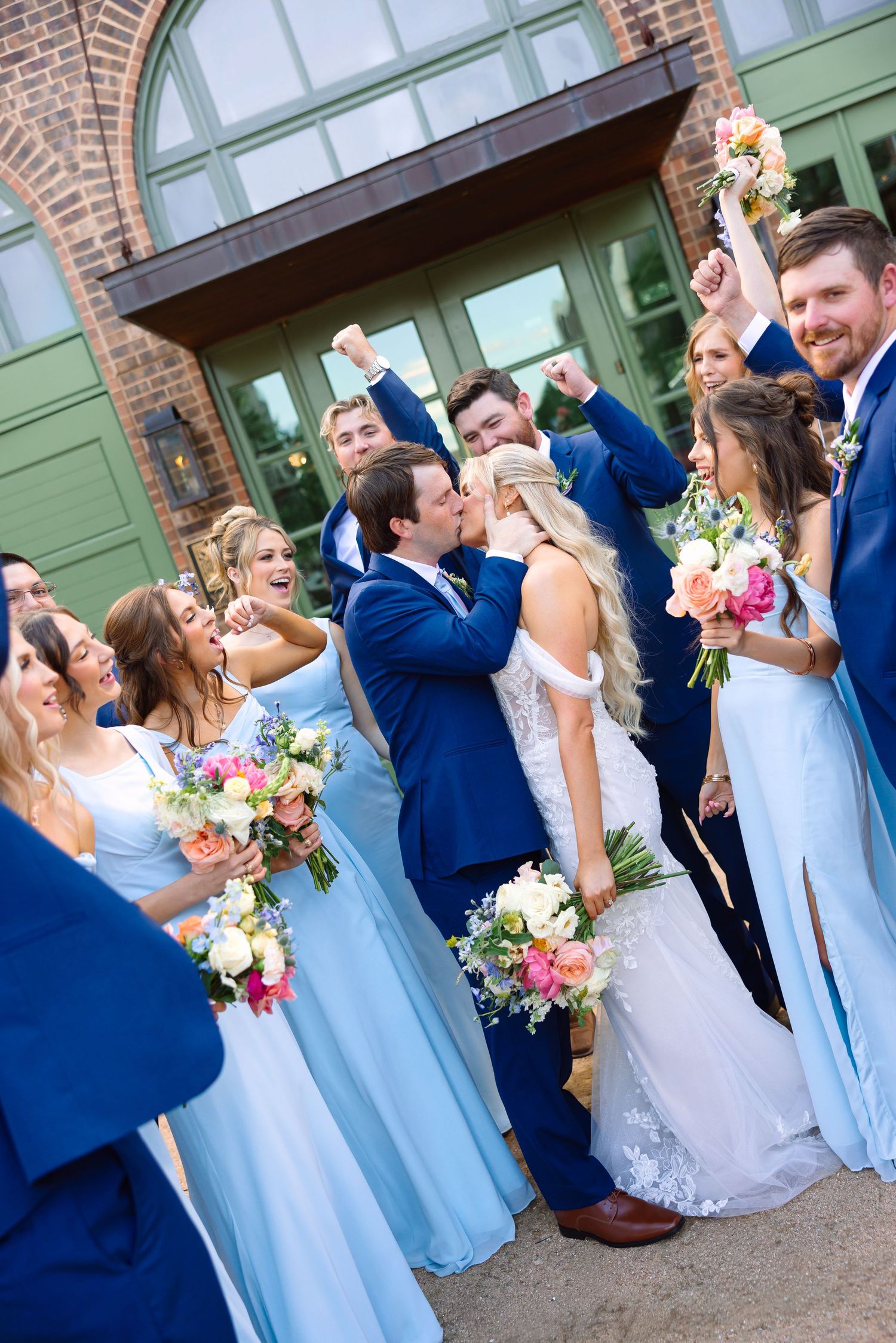 Bride and groom share a kiss surrounded by cheering wedding party in blue attire holding colorful bouquets outdoors.