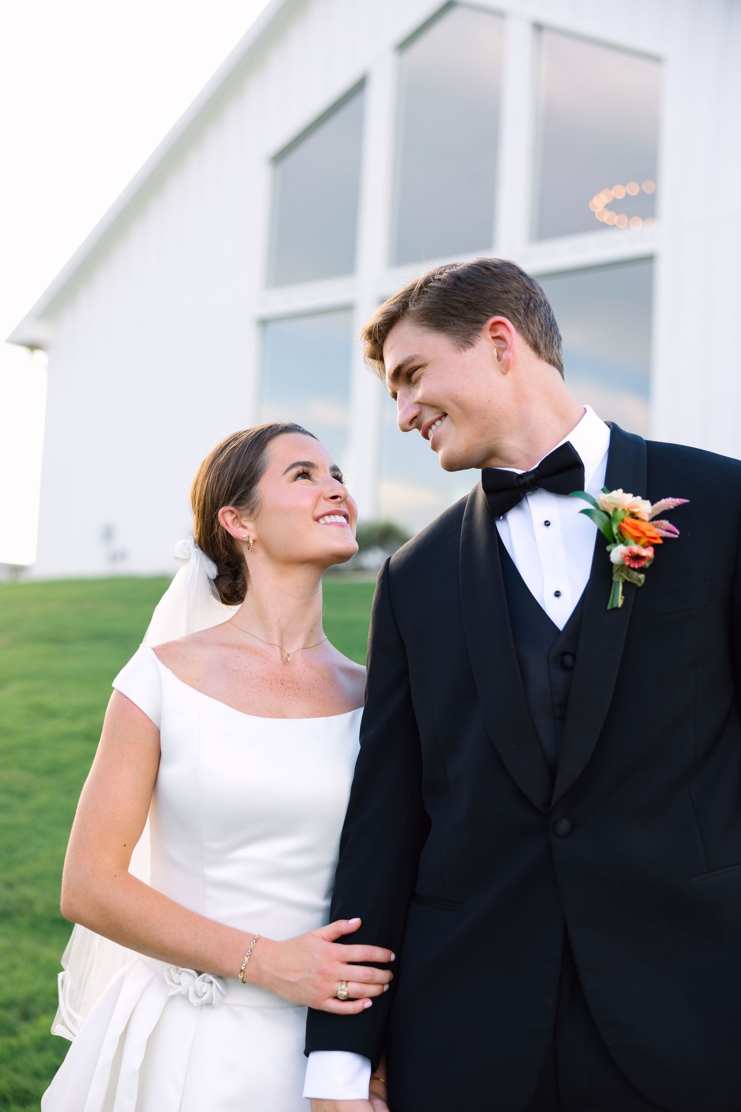 Bride and groom smiling at each other outdoors, she in white gown with veil, he in black tuxedo with floral boutonniere.