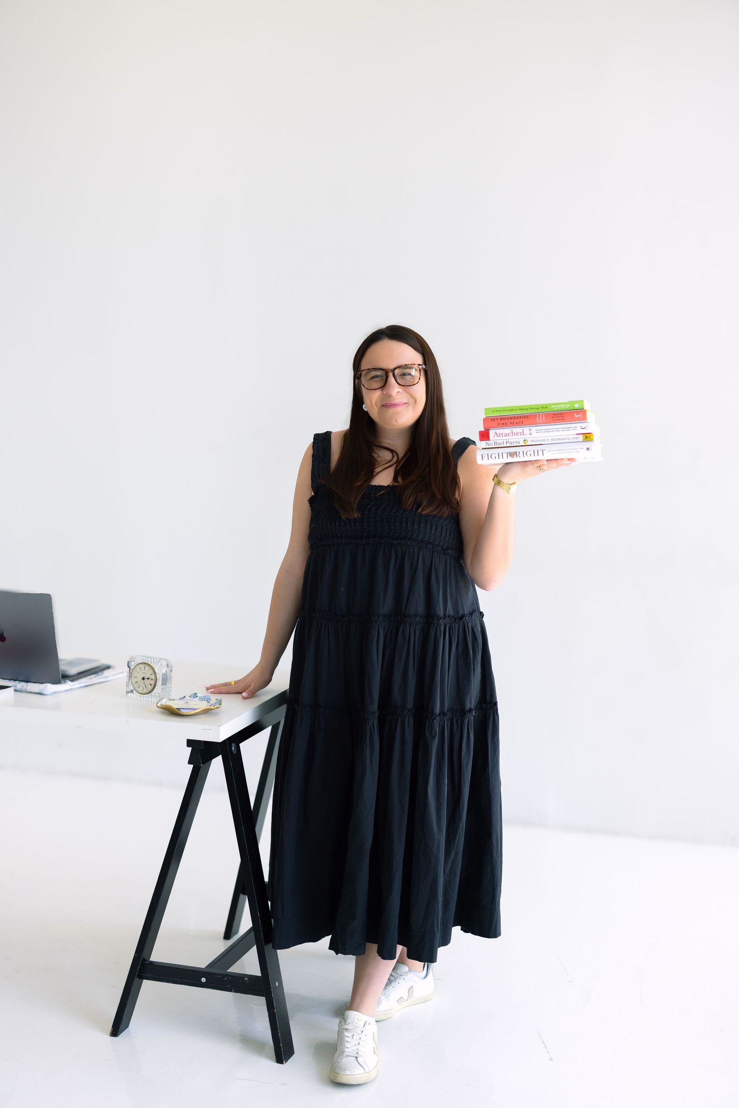 Woman in black dress holding stack of books beside a desk in a bright white minimalist office space.