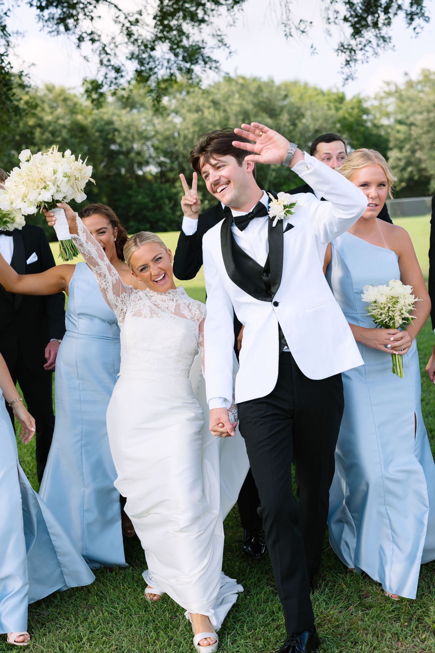 Joyful bride and groom in white tuxedo celebrate with wedding party in light blue dresses on green lawn.