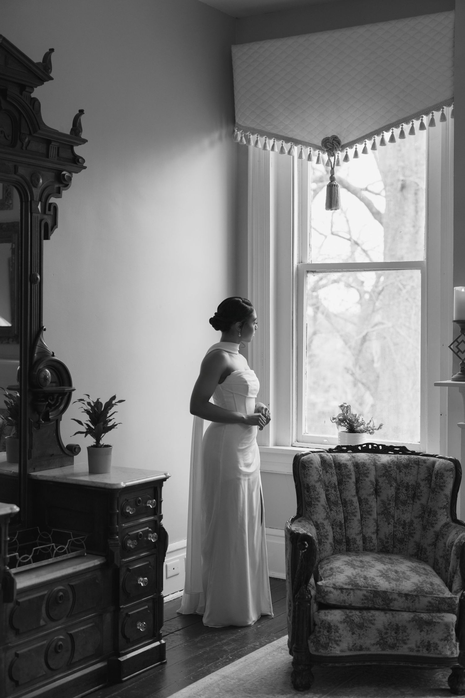 Black and white photo of a bride in a white gown standing by a vintage window, beside an ornate dresser and mirror.