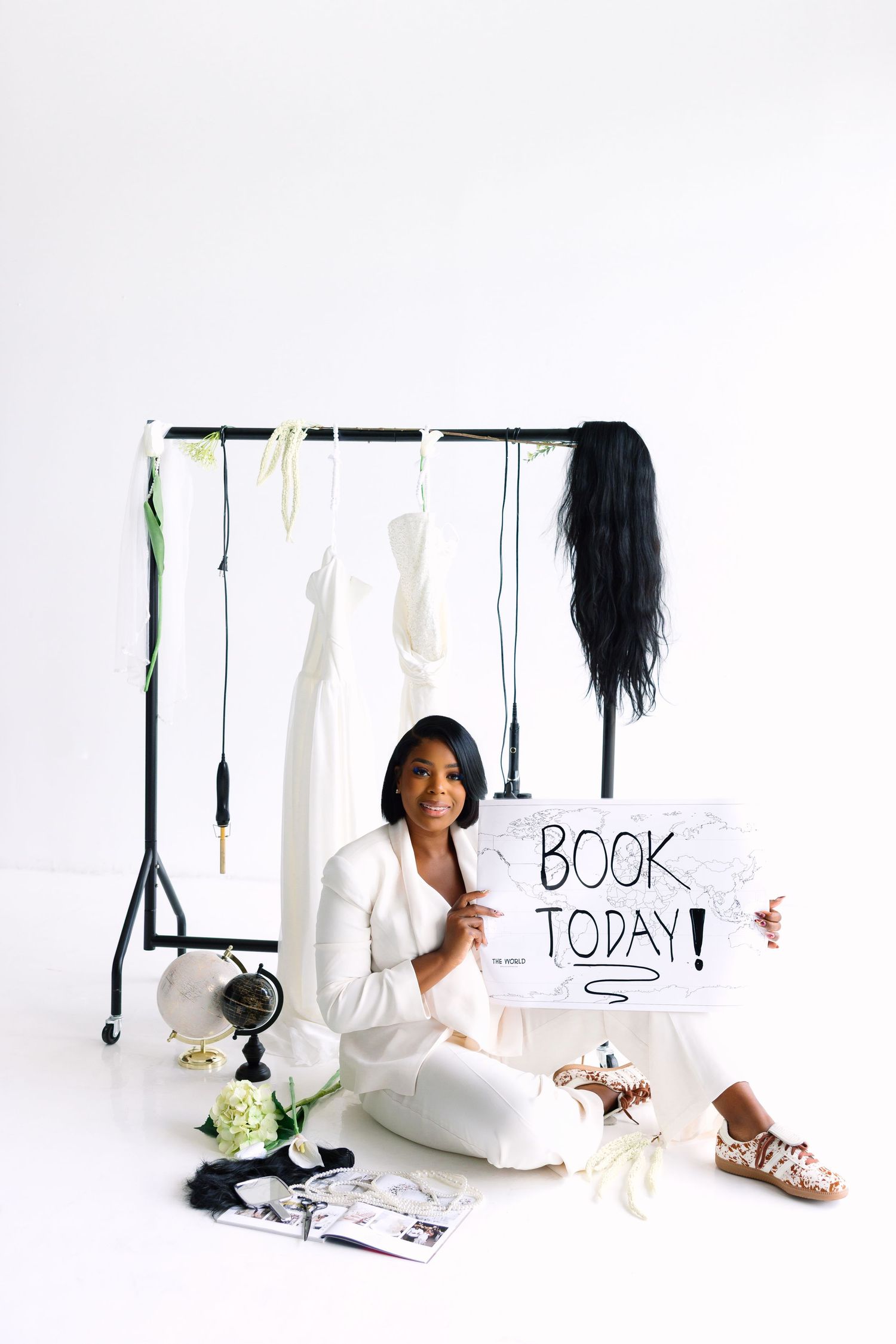 Woman in white suit sits on floor holding Book Today sign near clothing rack with dresses and accessories.