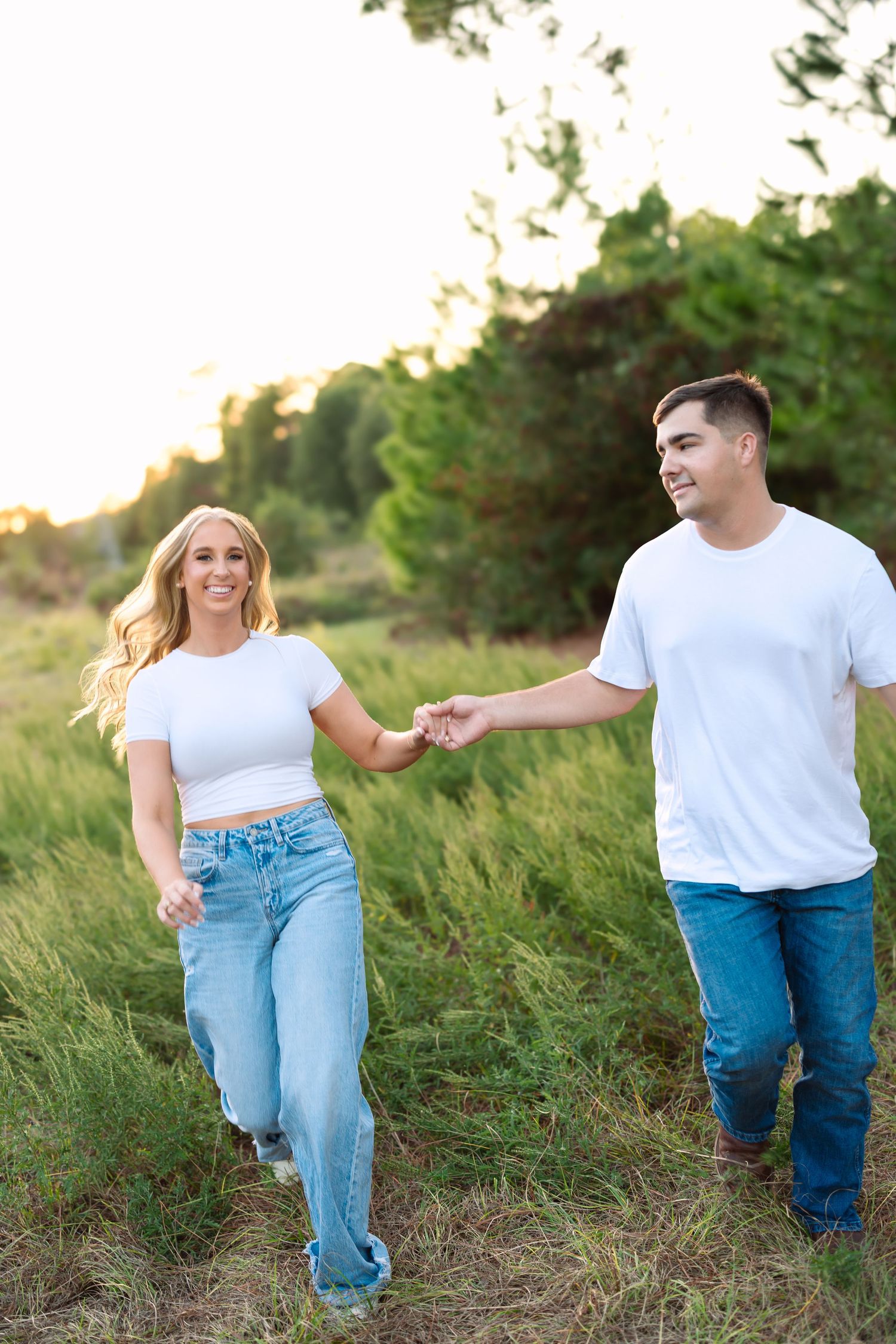 A smiling couple in matching white tops and jeans hold hands while walking through a lush green field at sunset.