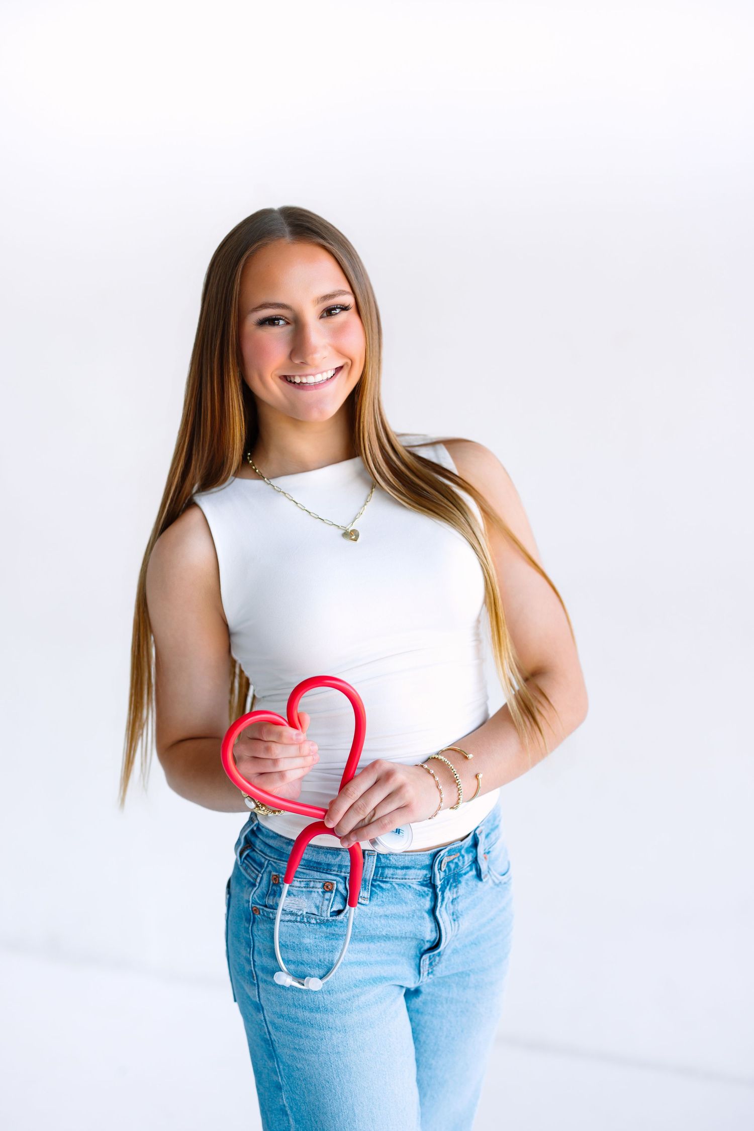 Casual lifestyle portrait against white background wearing white tank top and jeans while making heart shape with hands.