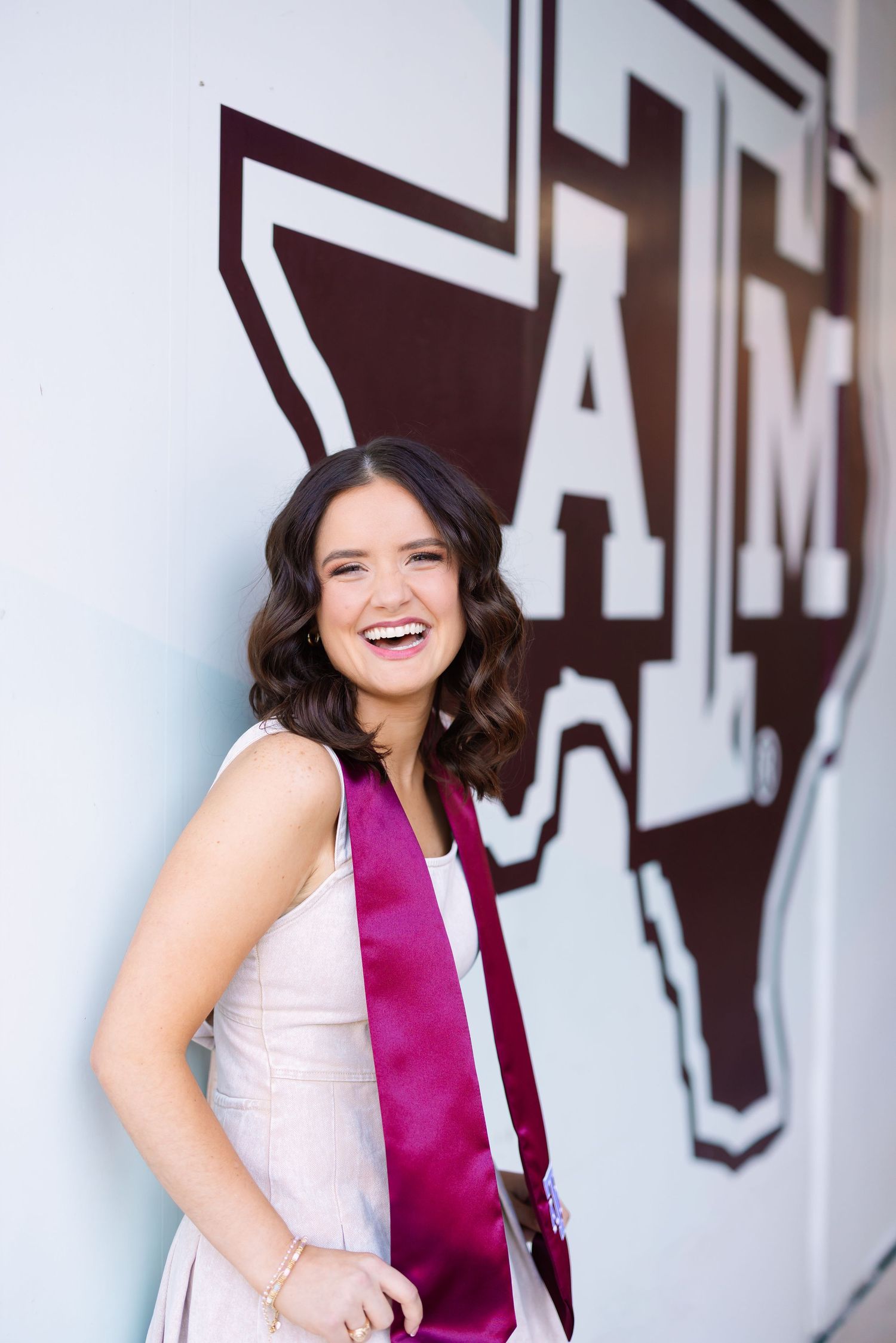 Graduate in pink dress poses with maroon stole against large Texas A&M logo wall.