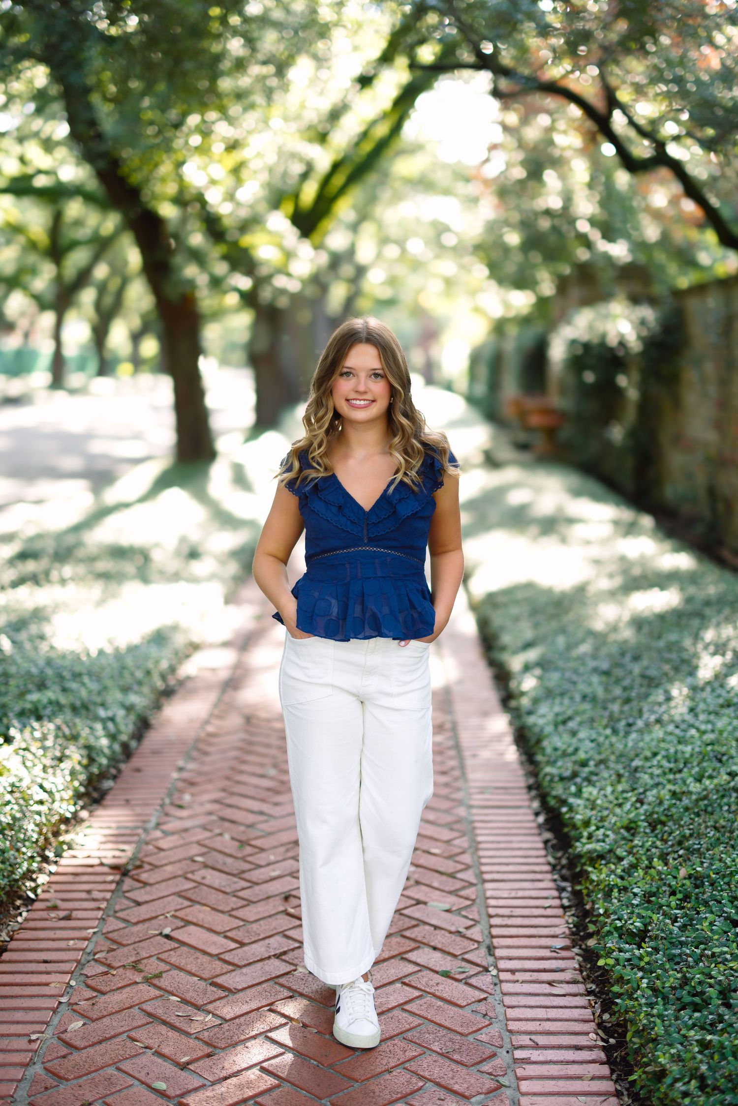 Young woman in blue top and white pants walks along a brick path lined with green hedges and trees.