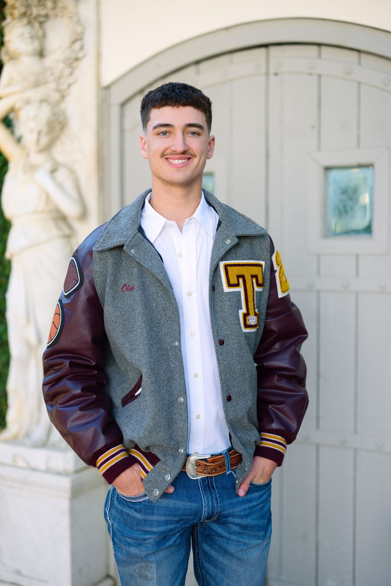 Young man smiling in a gray and maroon varsity letterman jacket with jeans, posing outdoors near a decorative door.