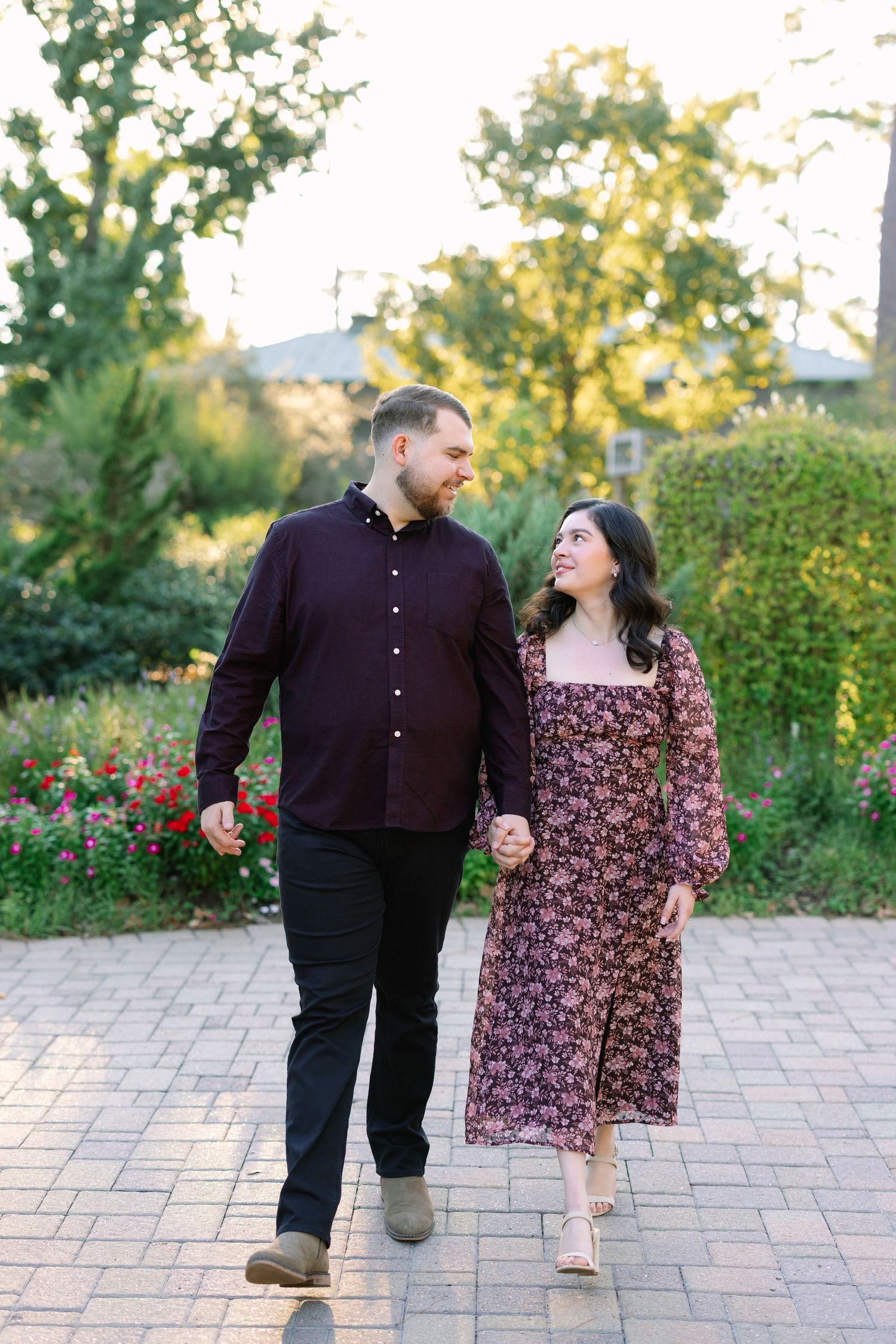Couple holding hands walking through a lush garden path, smiling at each other in coordinating burgundy outfits.