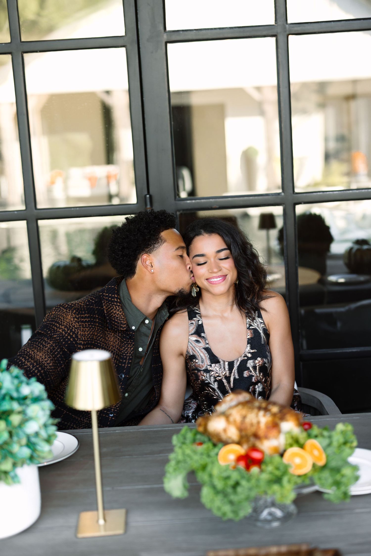 A couple shares a romantic kiss at a beautifully set dining table with fresh fruit and greenery centerpieces.
