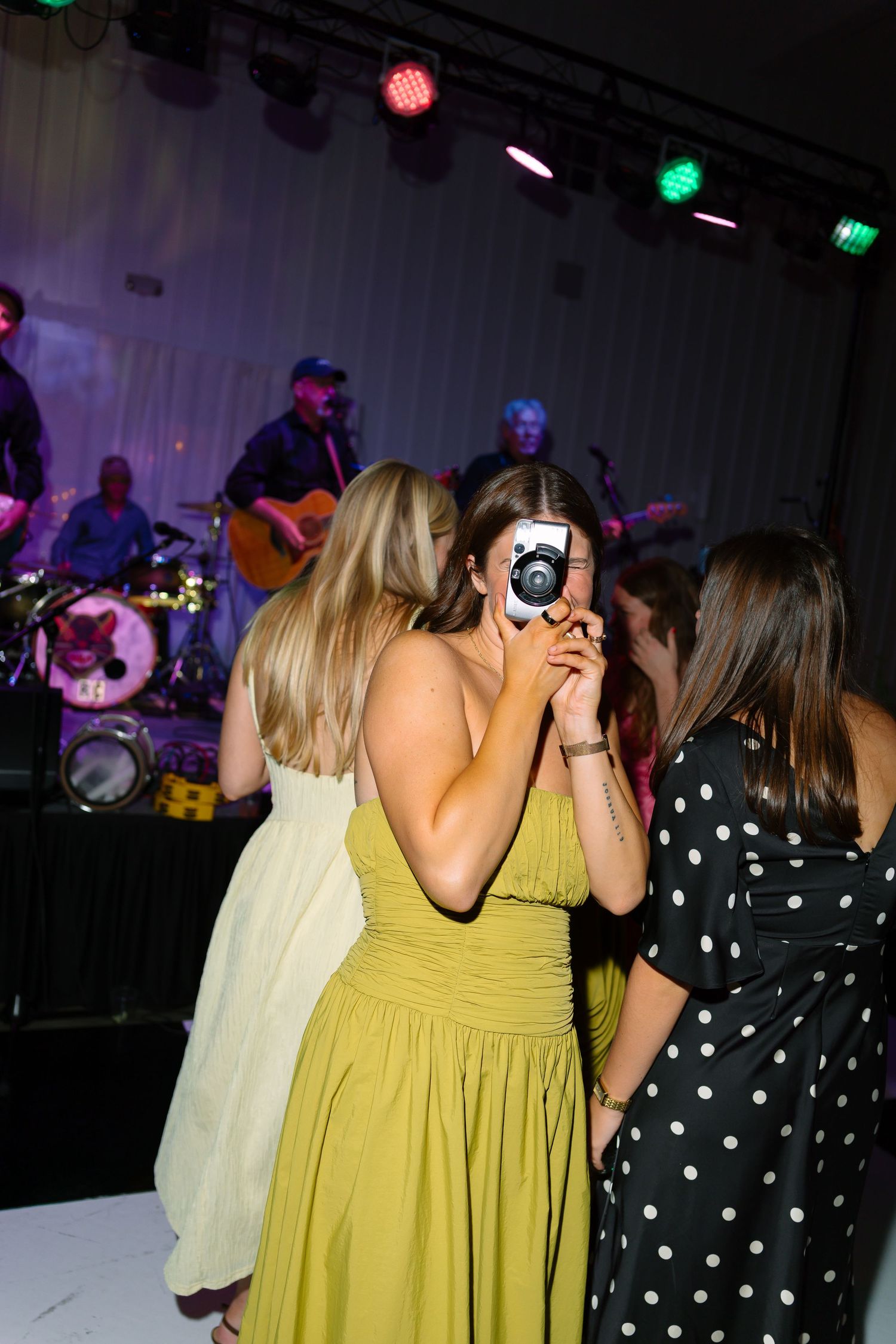 Women in colorful dresses taking photos with instant camera at live music event with band performing on stage.