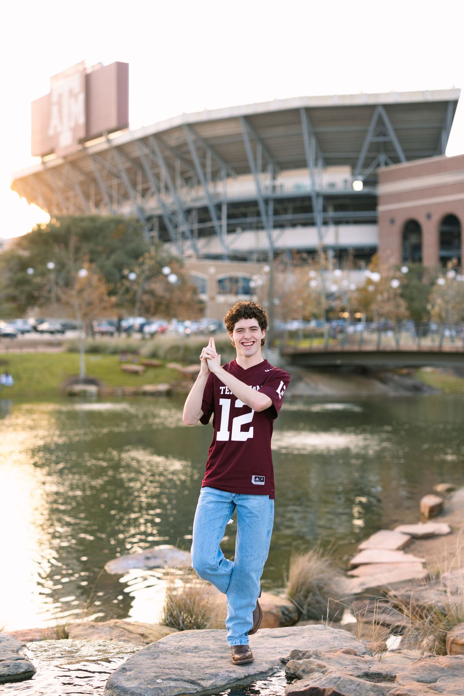 Student in maroon jersey poses on rocks near campus stadium and pond.