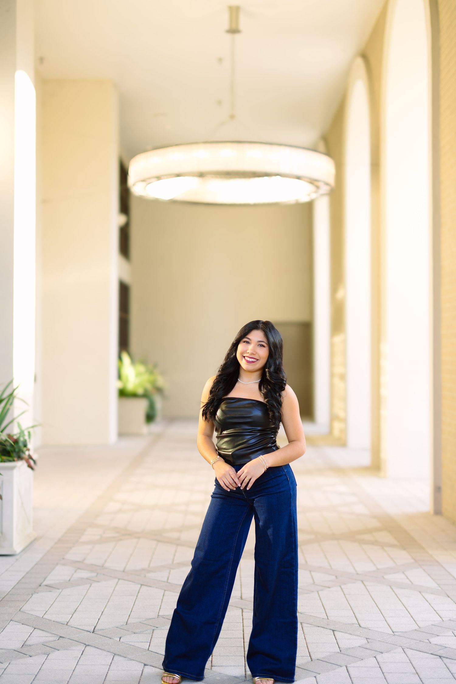 Woman in navy pants poses under circular light fixture in elegant hallway.