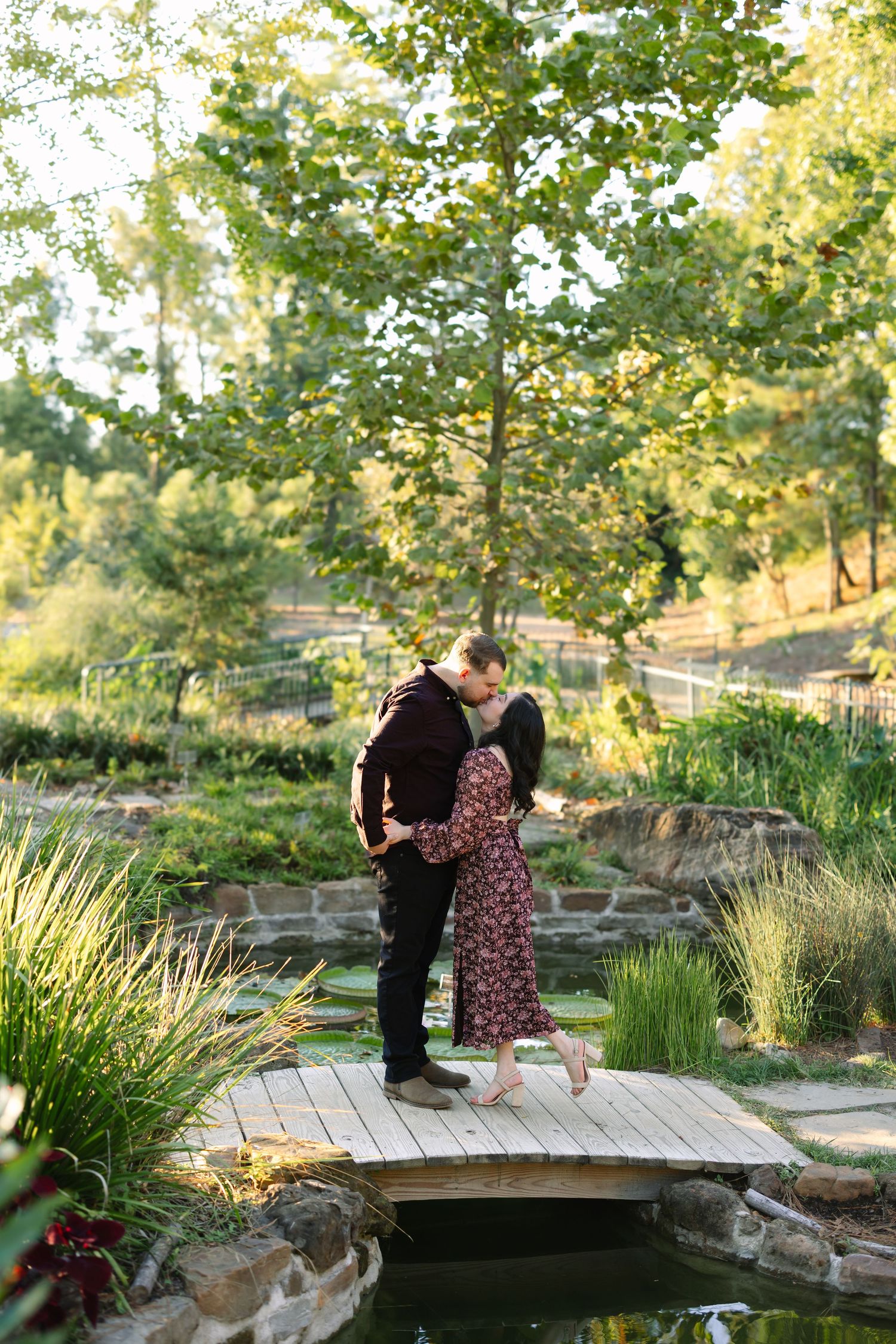 Couple sharing a kiss on a wooden bridge over a pond surrounded by lush greenery in a botanical garden.