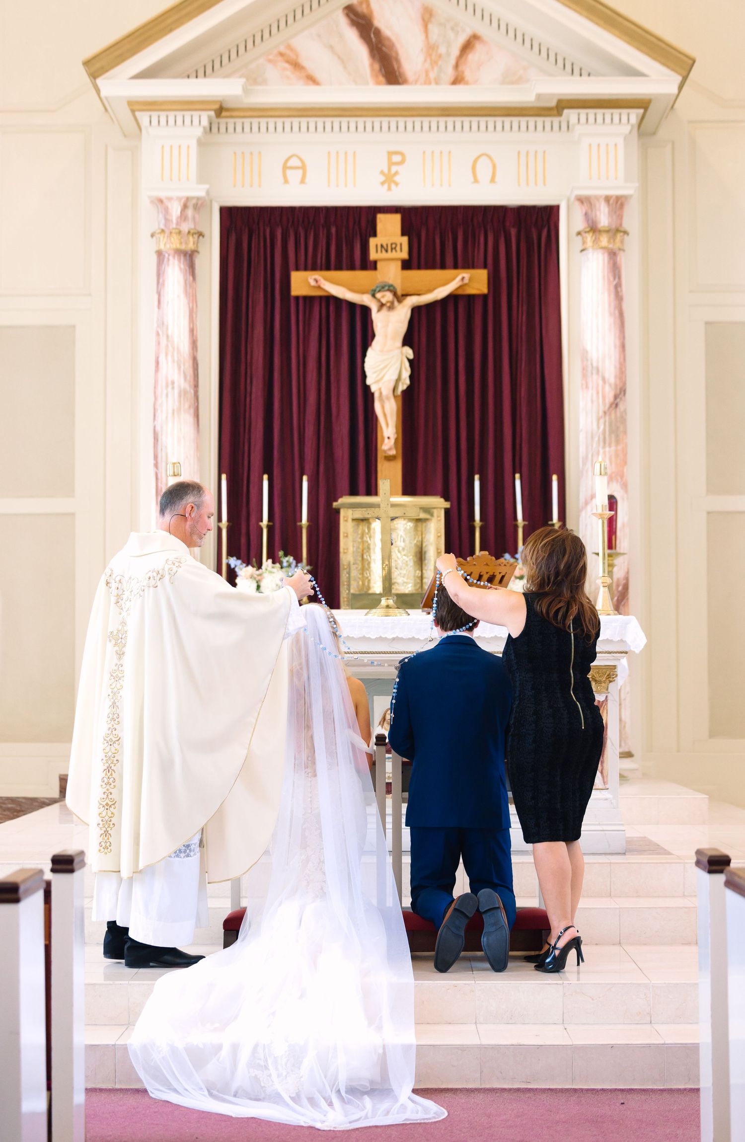 Bride and groom kneel before priest at ornate Catholic church altar during traditional wedding ceremony.