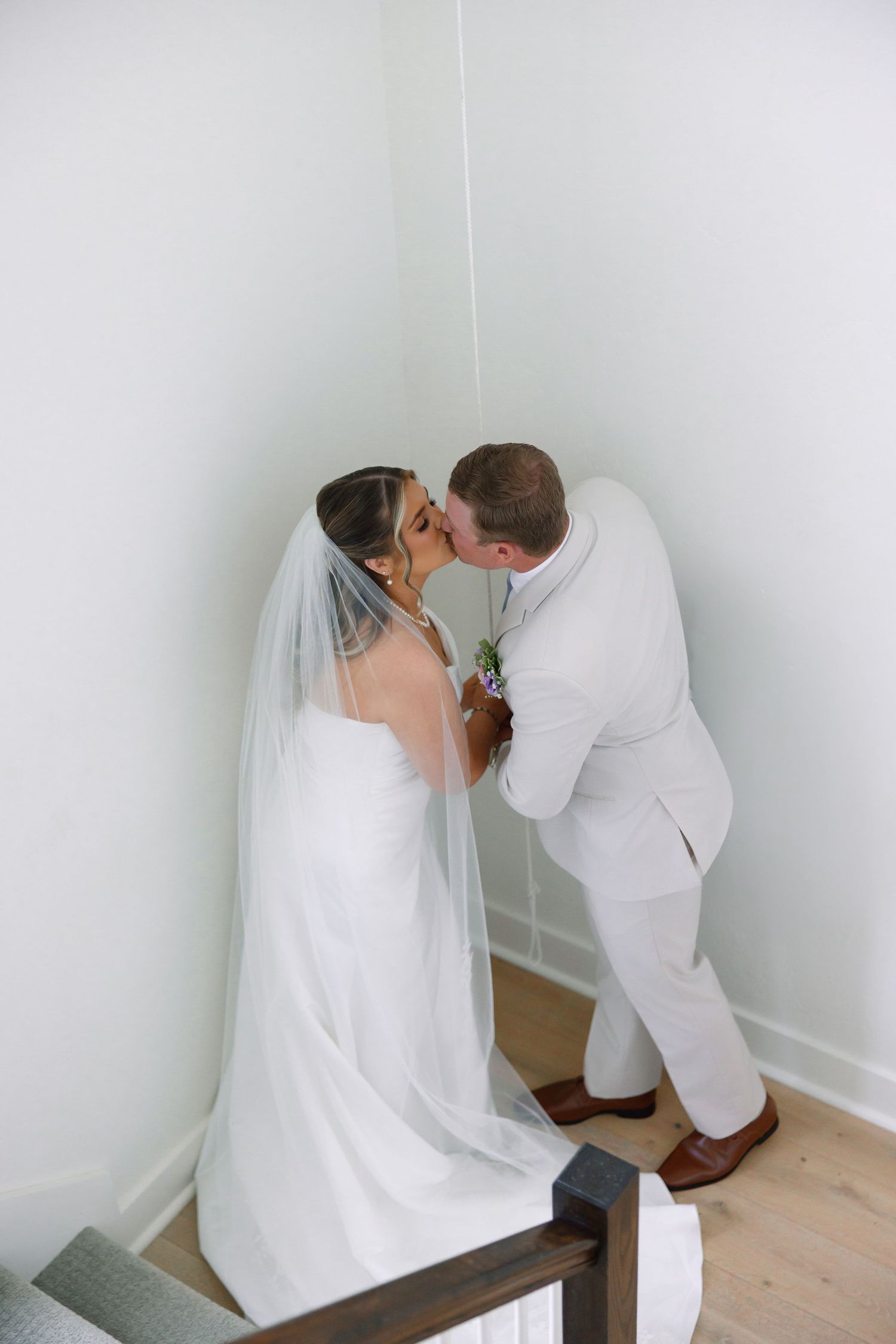 Bride and groom sharing a kiss in a white corner stairwell, bride wearing a flowing veil and white gown.
