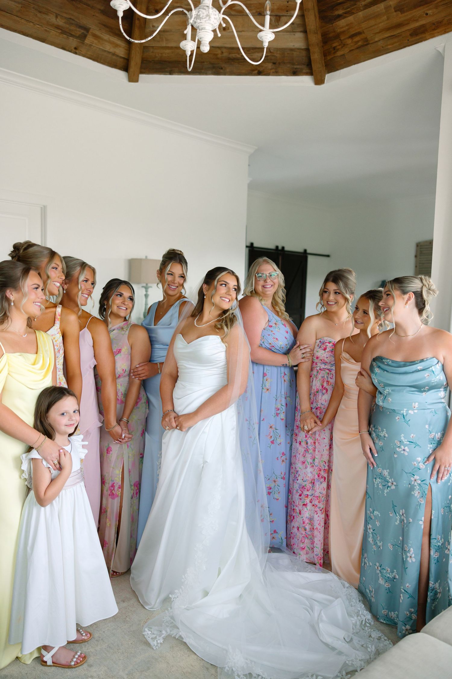 Bride in white gown poses with bridesmaids in pastel mismatched dresses inside an elegant venue.