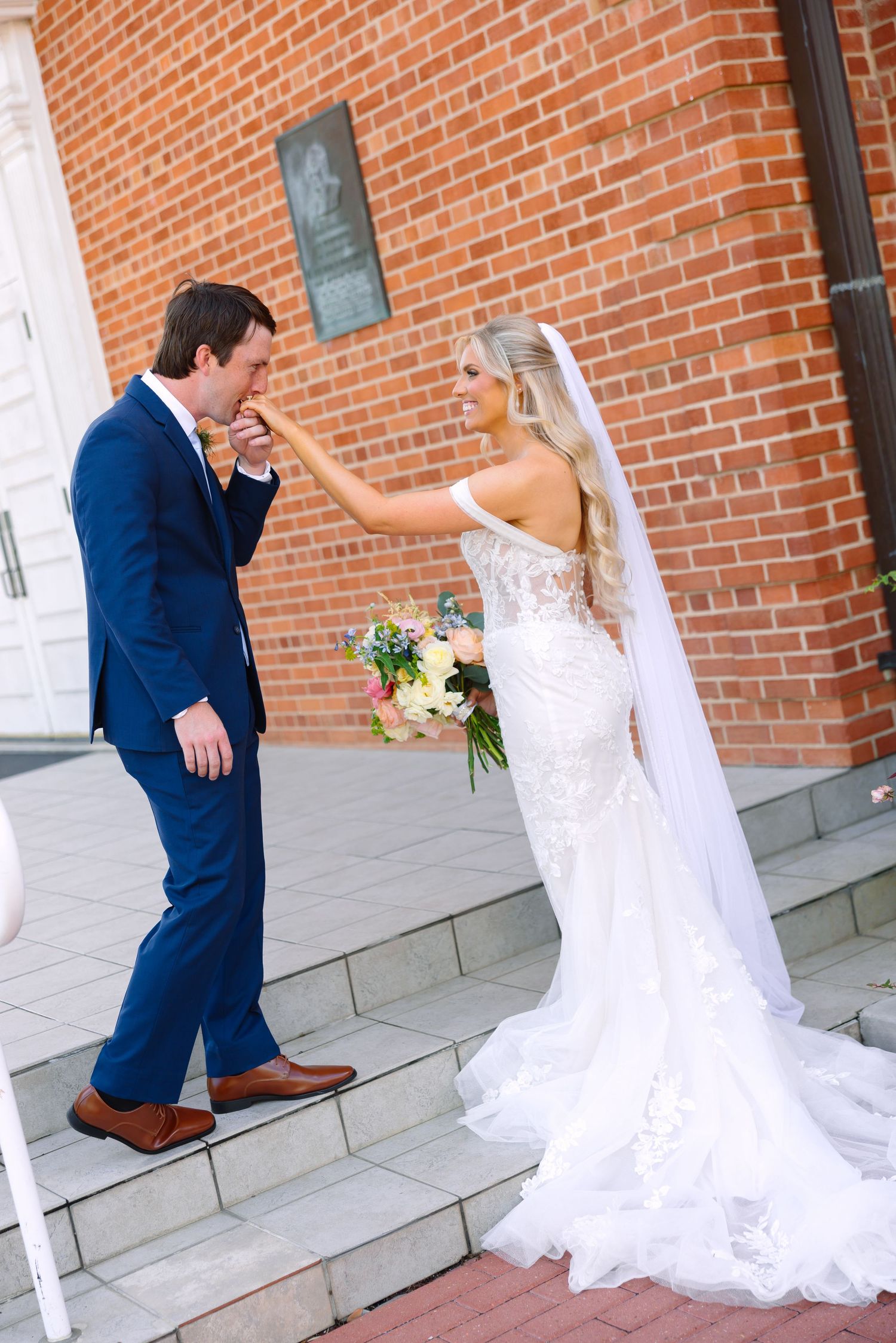 Groom in navy suit kisses bride's hand on brick church steps during outdoor wedding portrait.