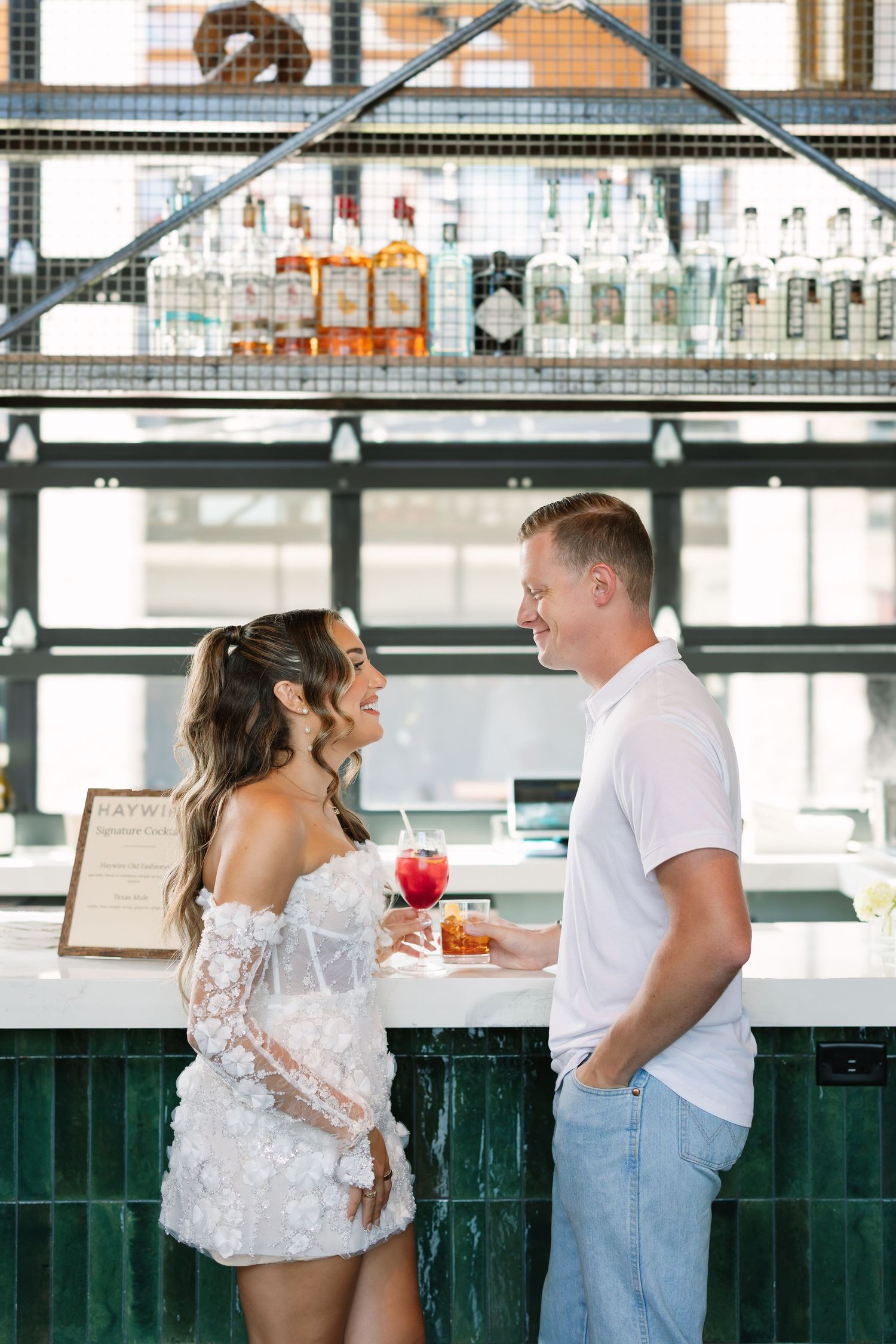 Bride in white lace mini dress laughing with groom at elegant bar during wedding celebration.