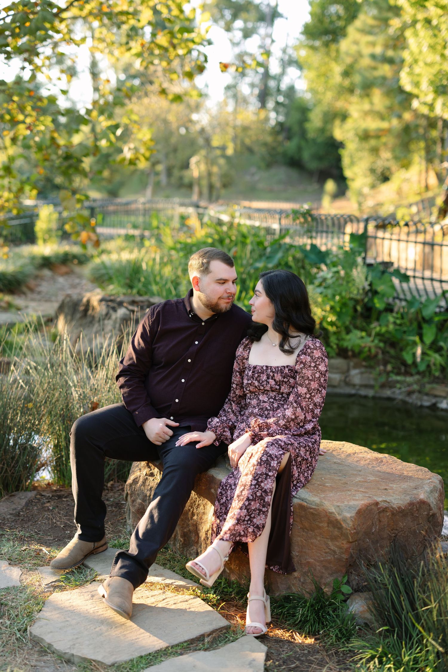 Couple sitting close together on a large rock in a sunlit garden, gazing into each other's eyes.
