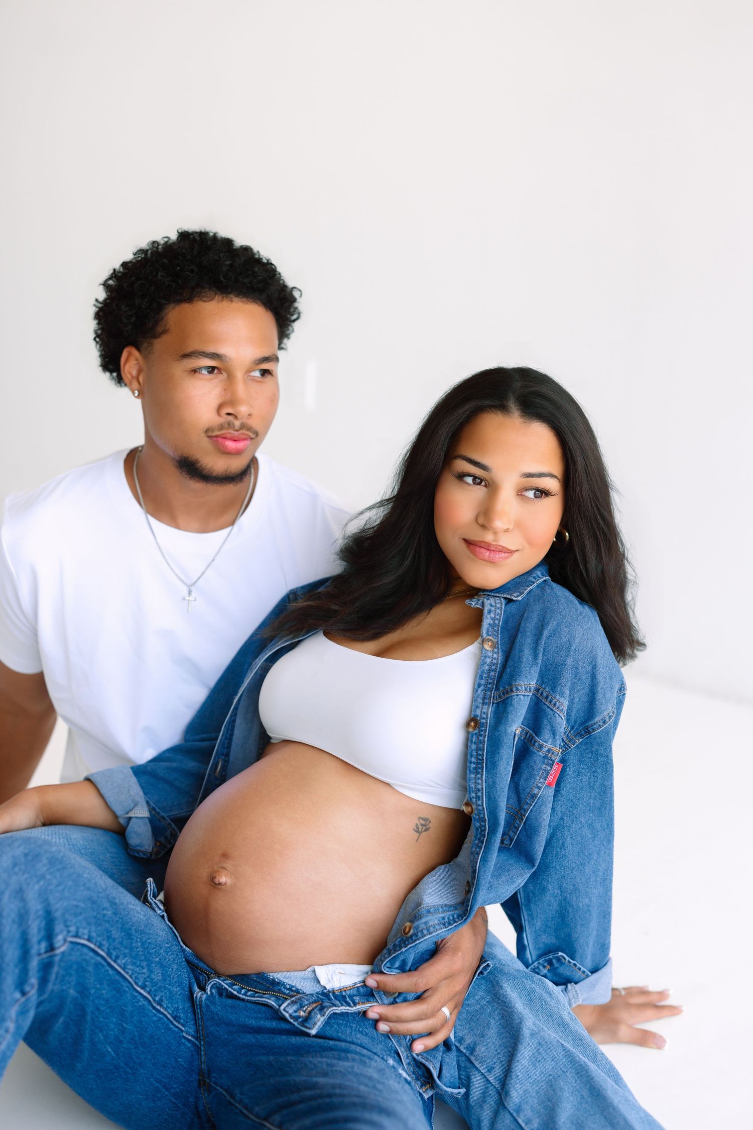 A pregnant couple in denim and white poses together, the woman cradling her baby bump in a bright studio setting.