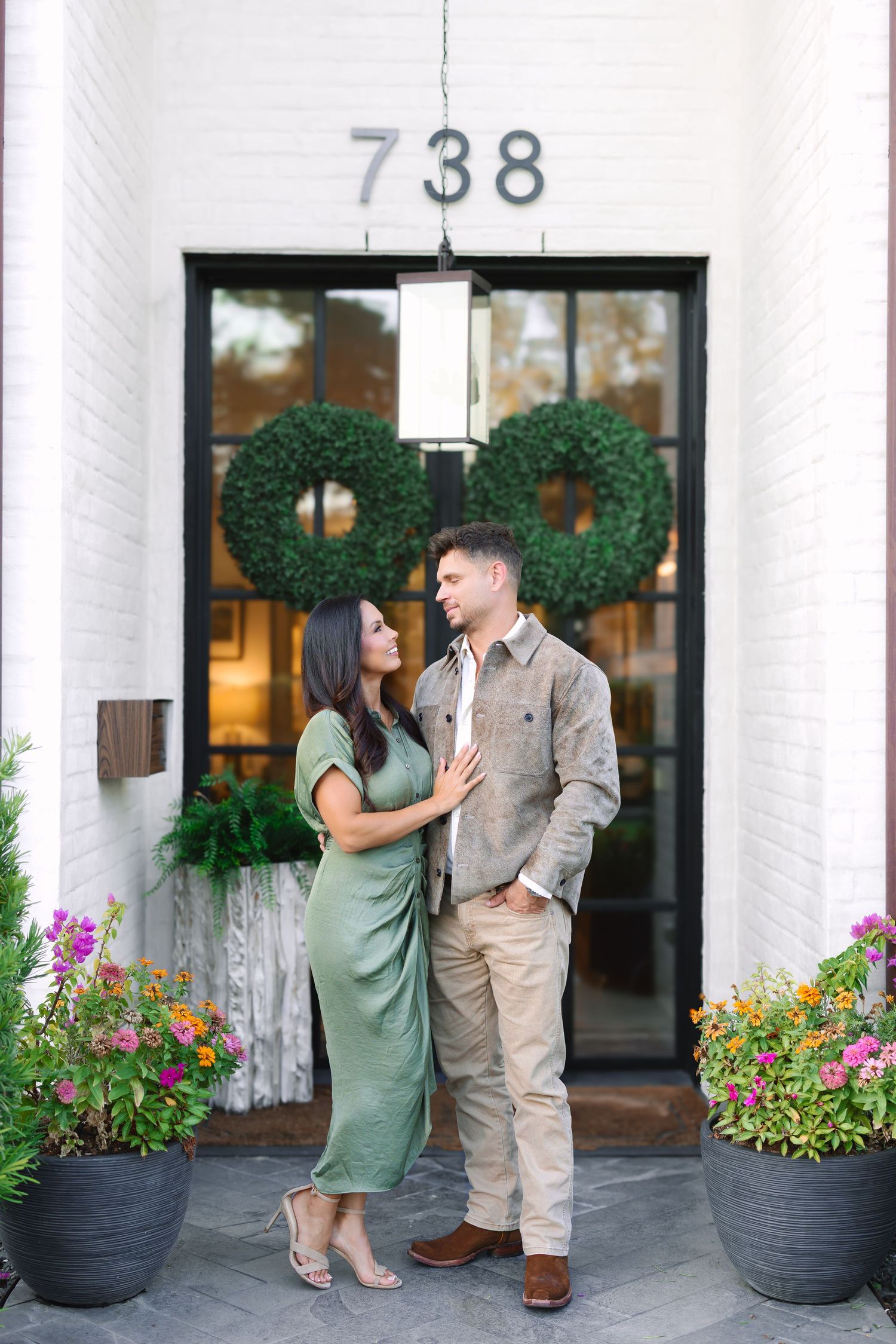 Couple in green dress and khaki outfit standing together in front of white brick building with wreaths and flowers.