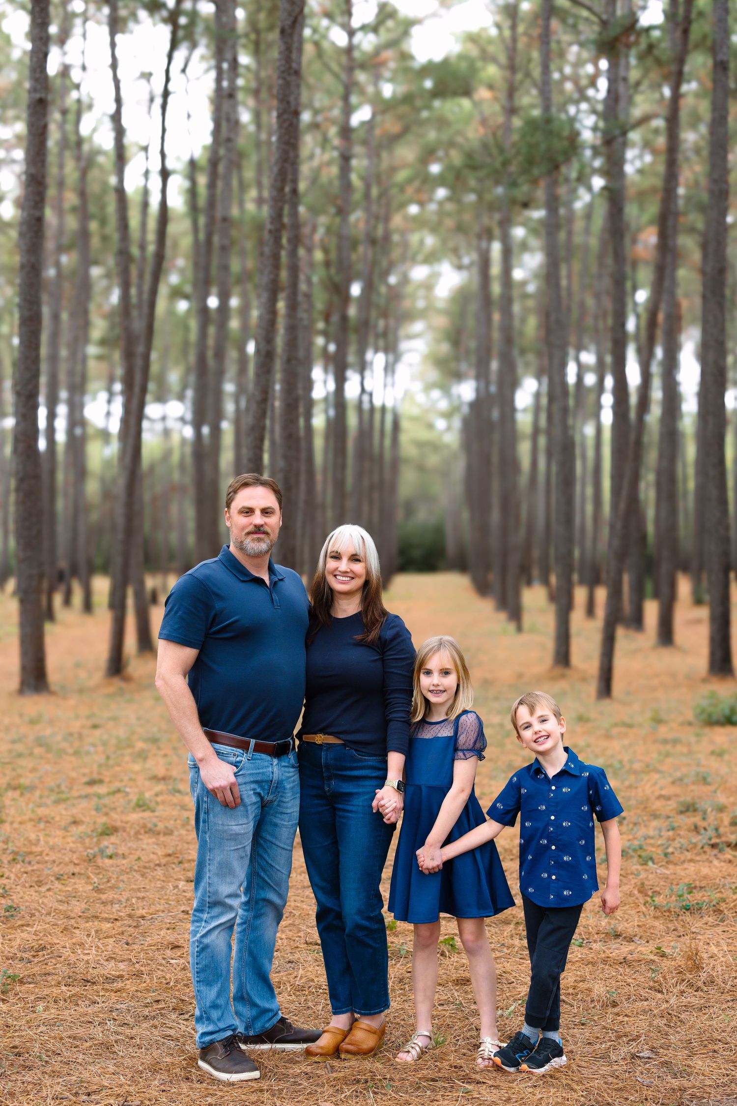 A family of four dressed in blue poses together in a pine forest for a portrait.