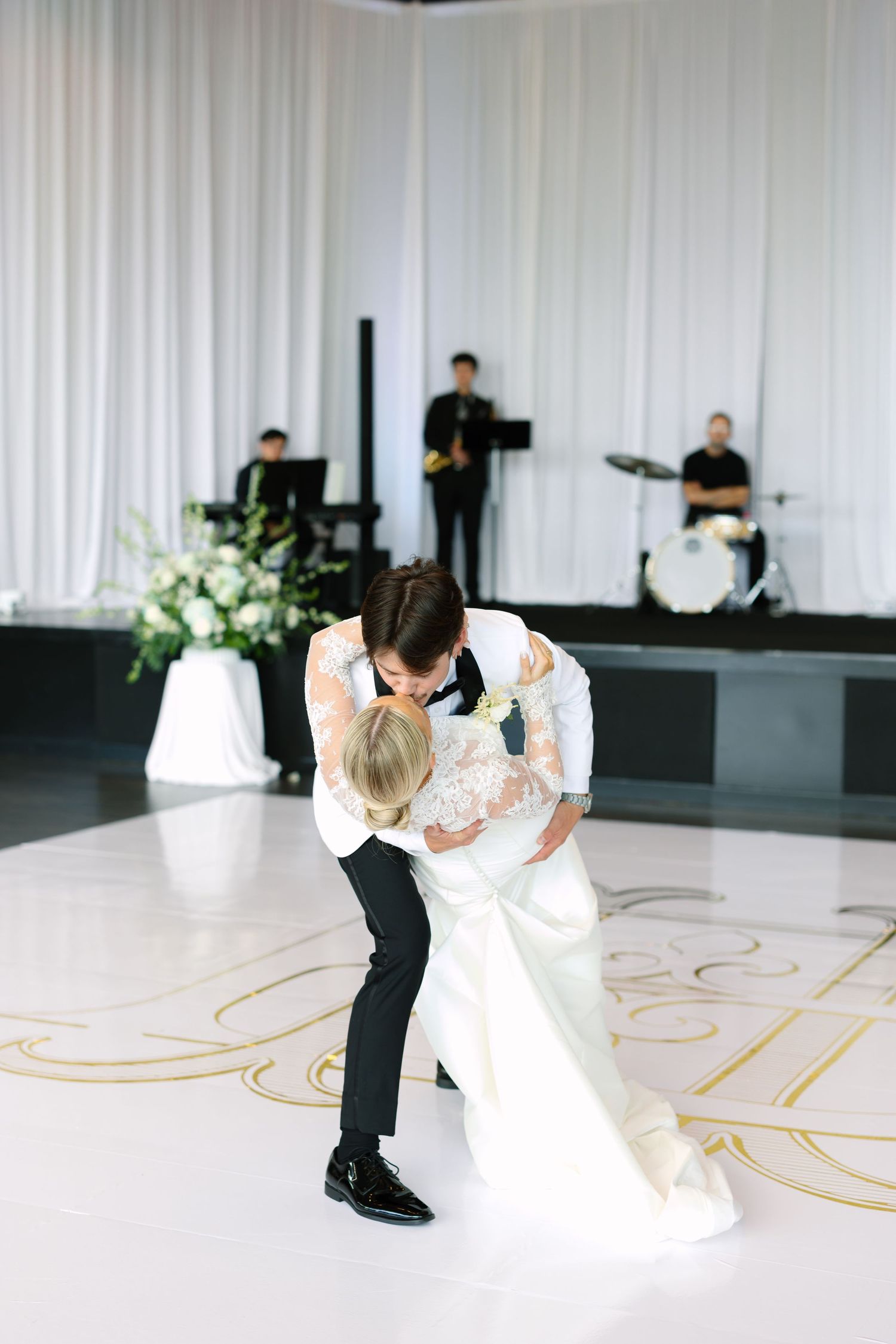 Bride and groom share romantic first dance dip on white dance floor with live band performing in elegant ballroom.