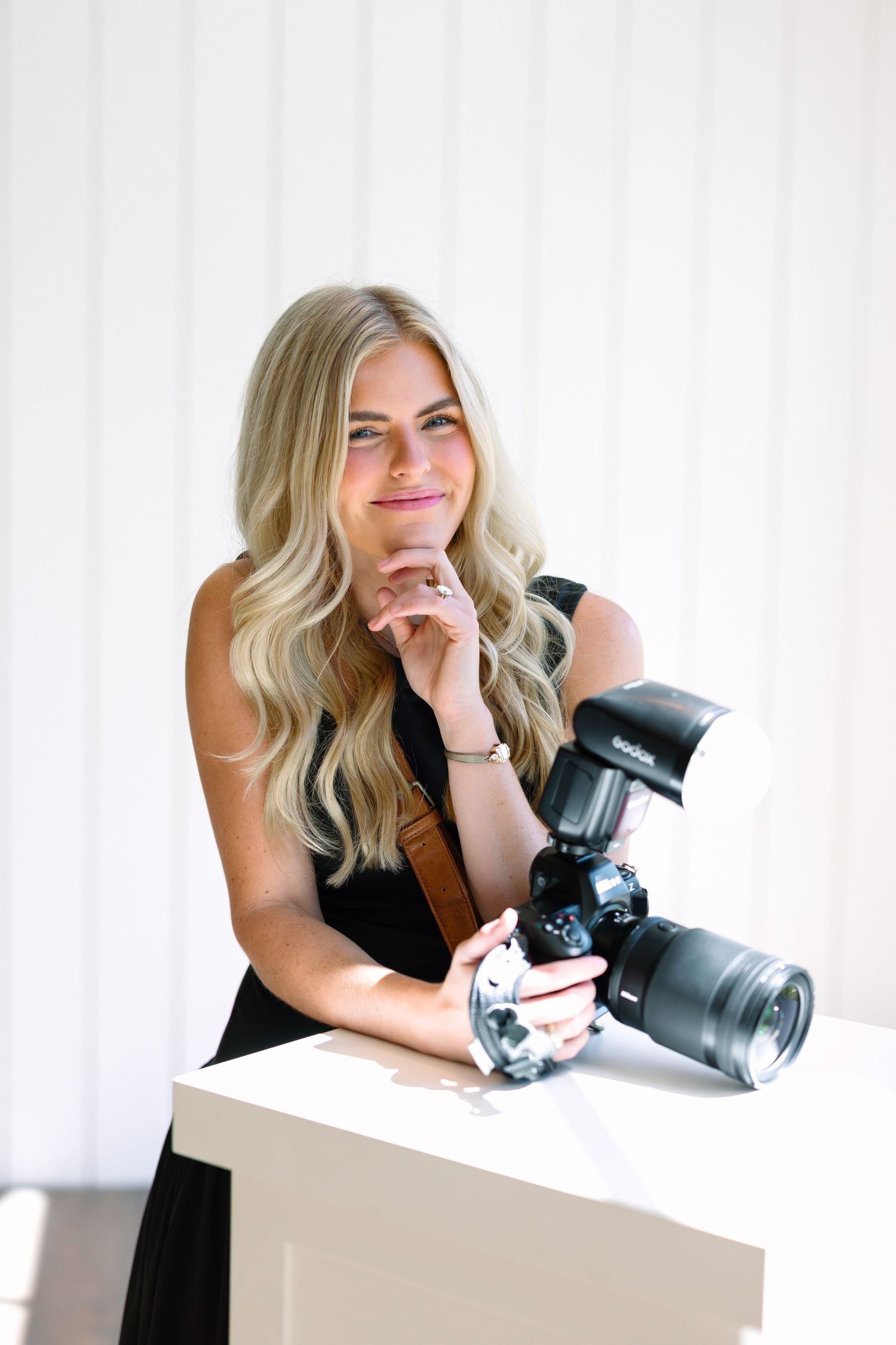 Blonde female photographer smiling and posing with a DSLR camera and flash on a white table.