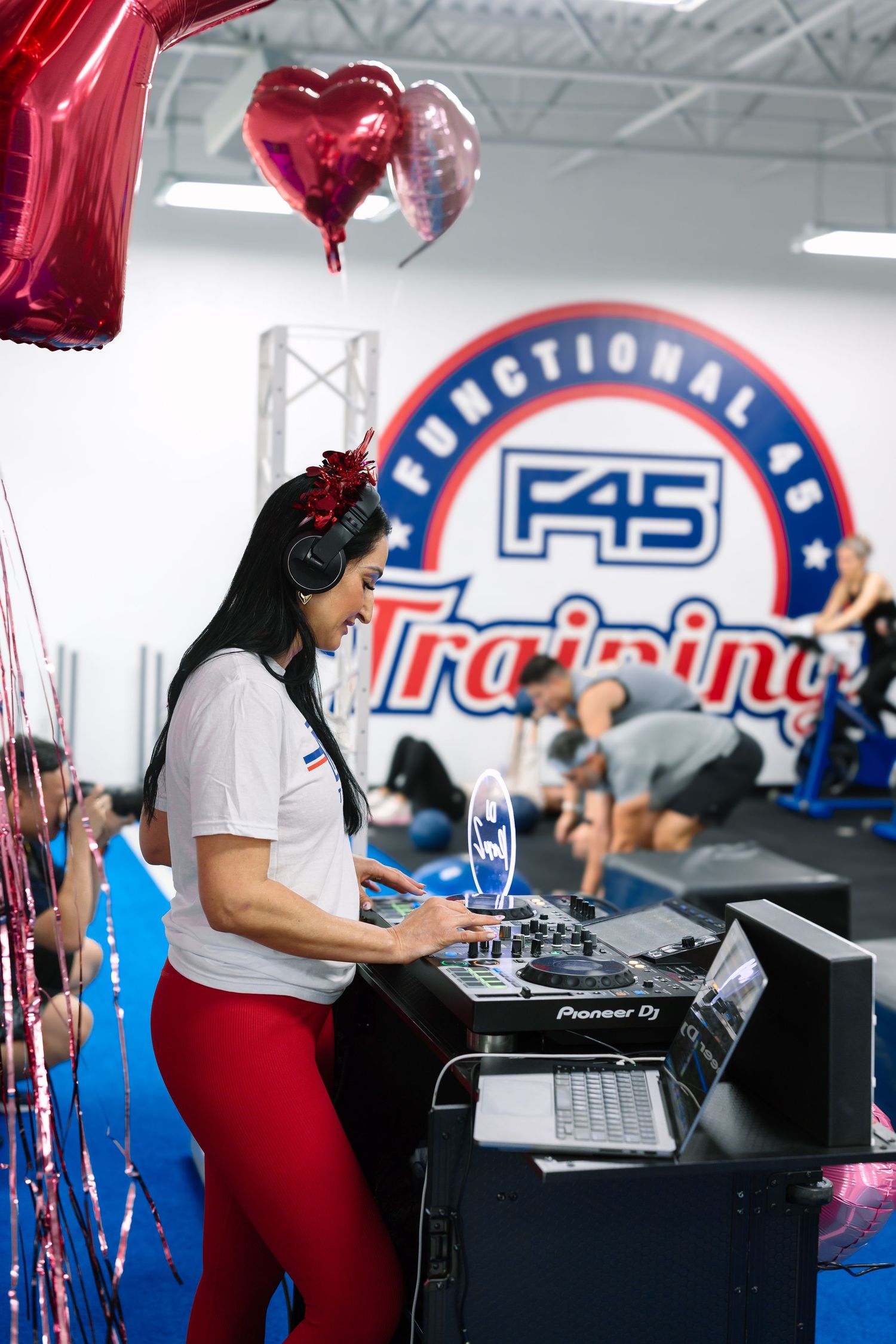 A female DJ in red pants and headphones operates a Pioneer DJ setup at an F45 Training gym event with heart balloons.