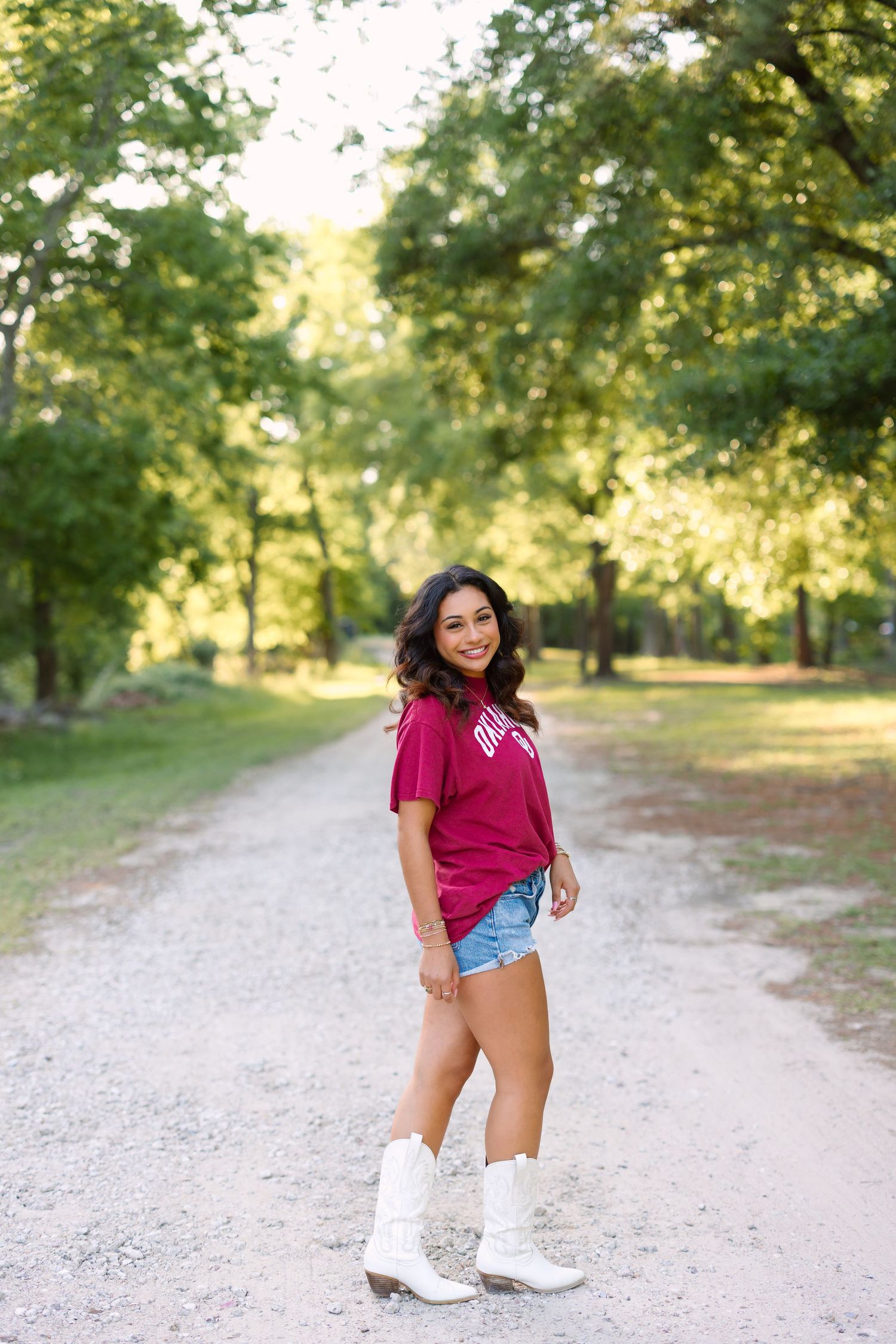 A person in a burgundy top and denim shorts poses on a sunlit gravel path surrounded by green trees.