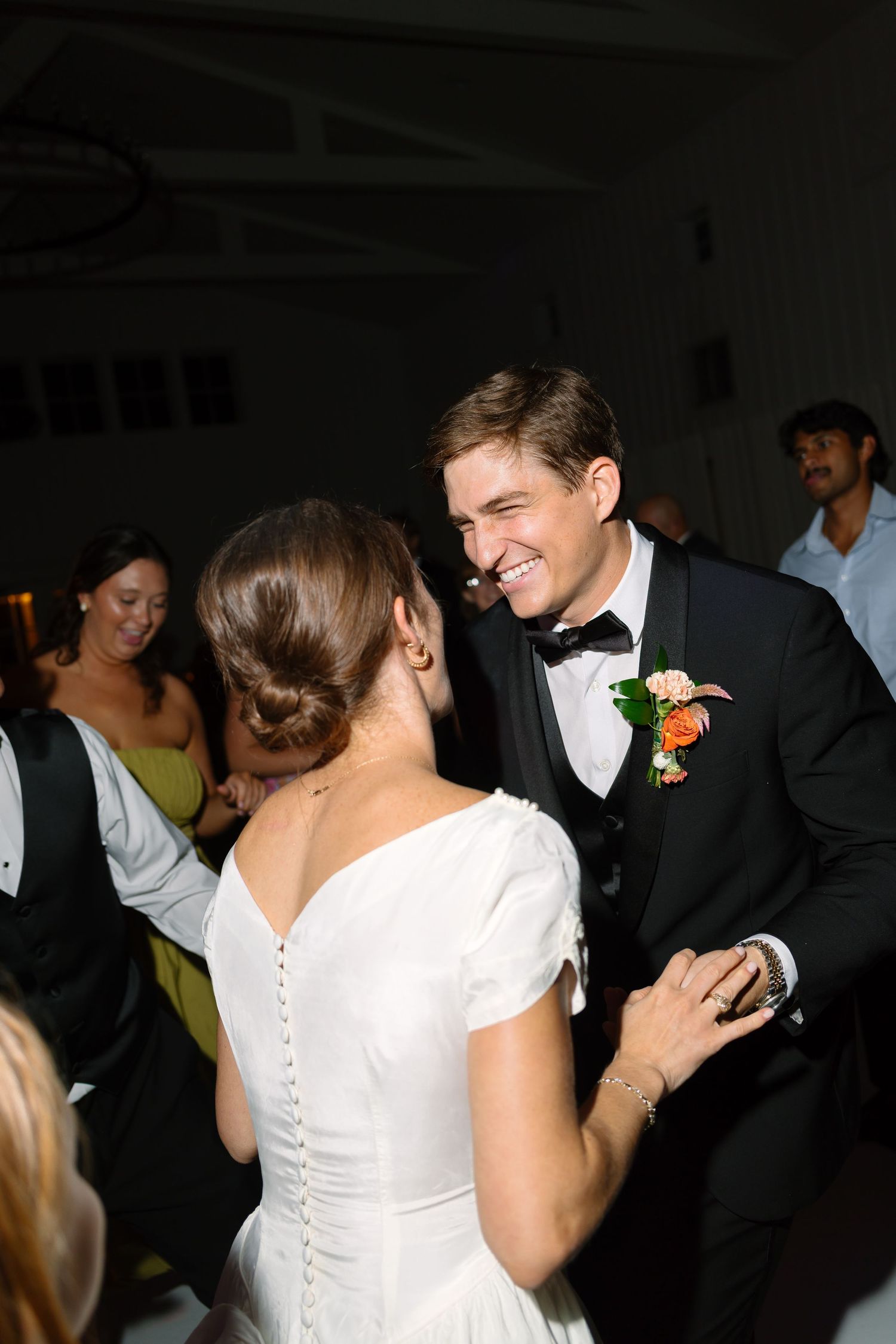 Bride and groom share a joyful first dance at their wedding reception, groom smiling in a black tuxedo with floral boutonniere.