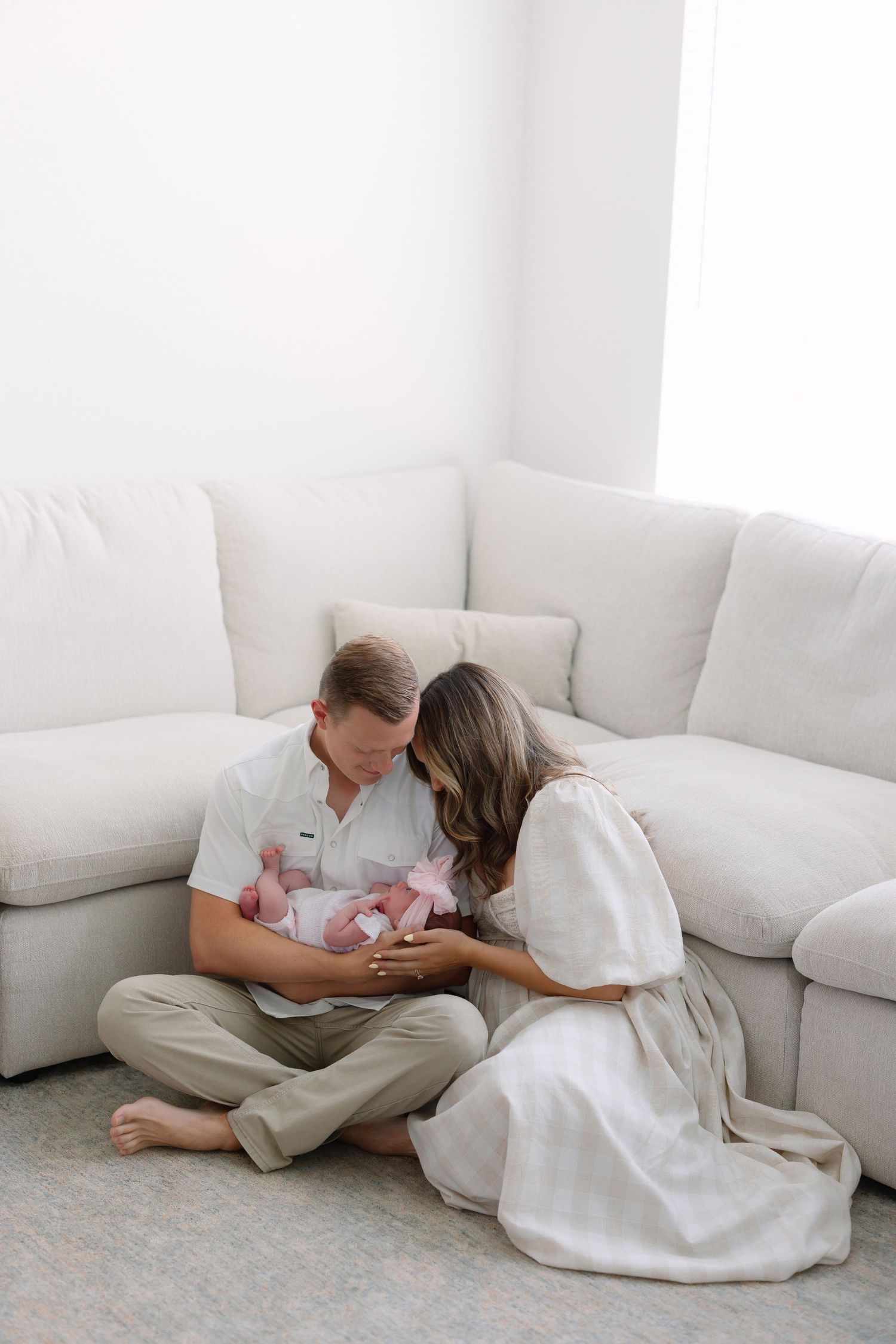 A couple sits on the floor against a white sofa, lovingly gazing at their newborn baby dressed in pink.