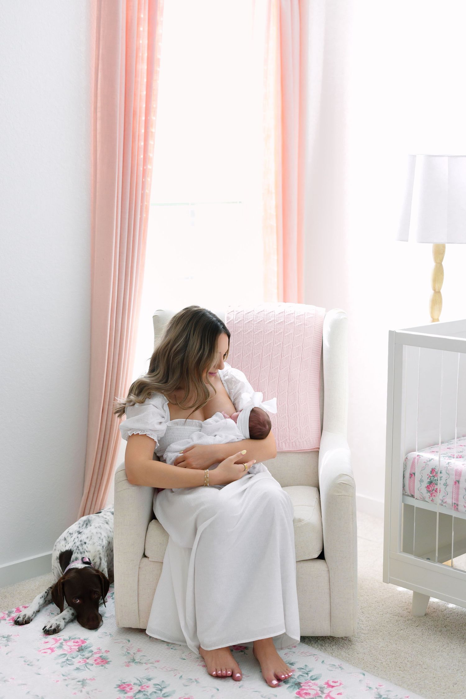 Mother in white dress breastfeeding newborn in nursery glider chair with dog beside her and pink curtains.
