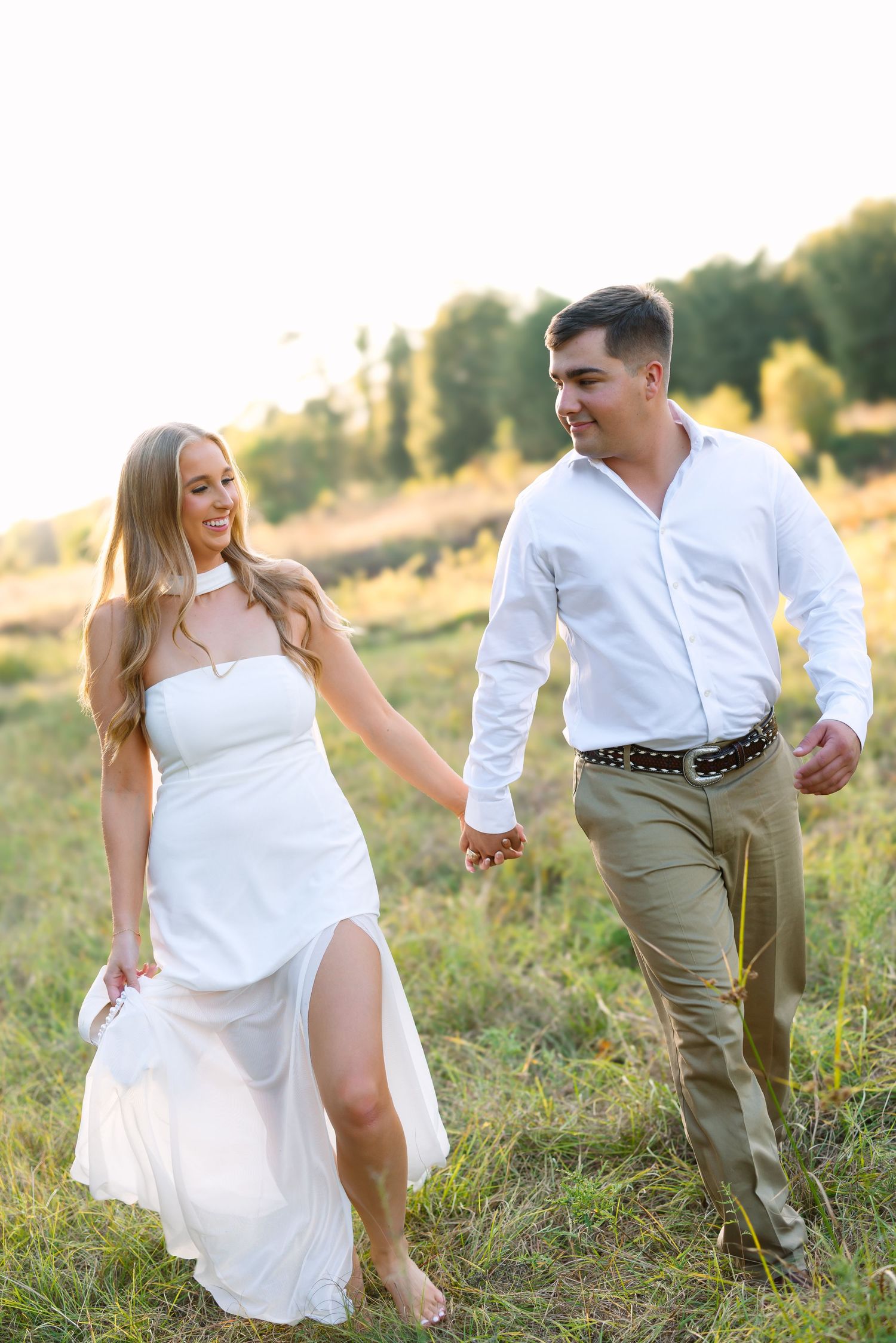 Couple in white and khaki outfits playfully running through a sunlit grassy field at golden hour.