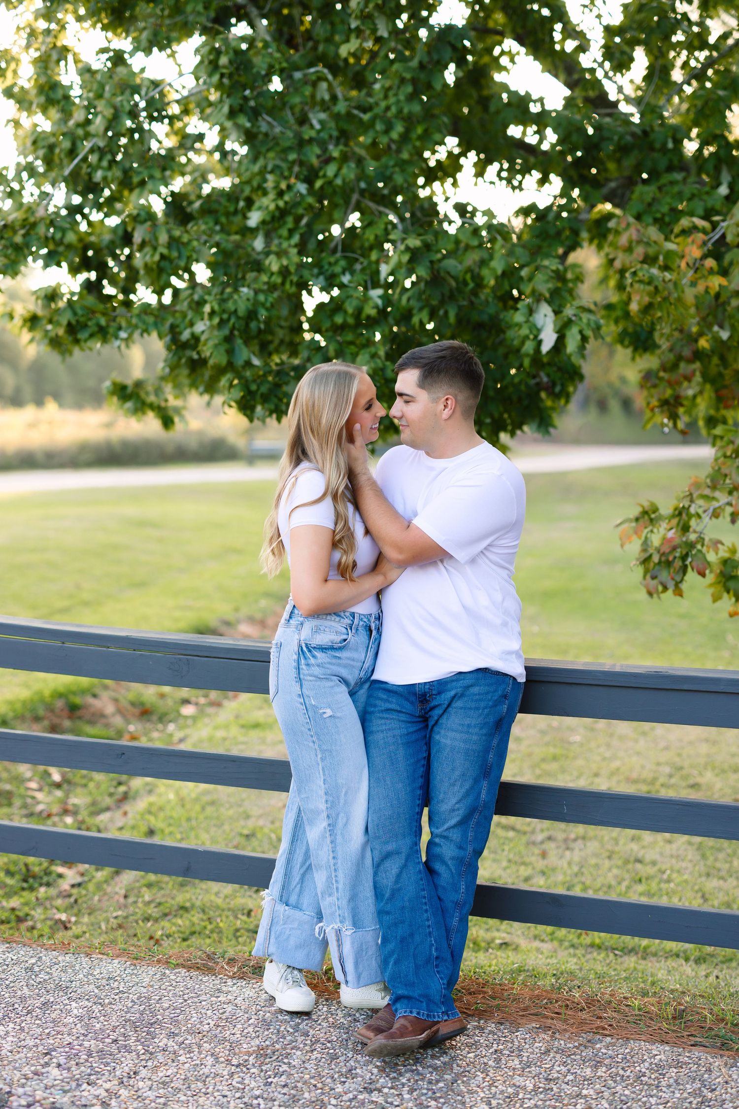Couple in matching white tops and jeans embracing by a wooden fence surrounded by lush green trees outdoors.